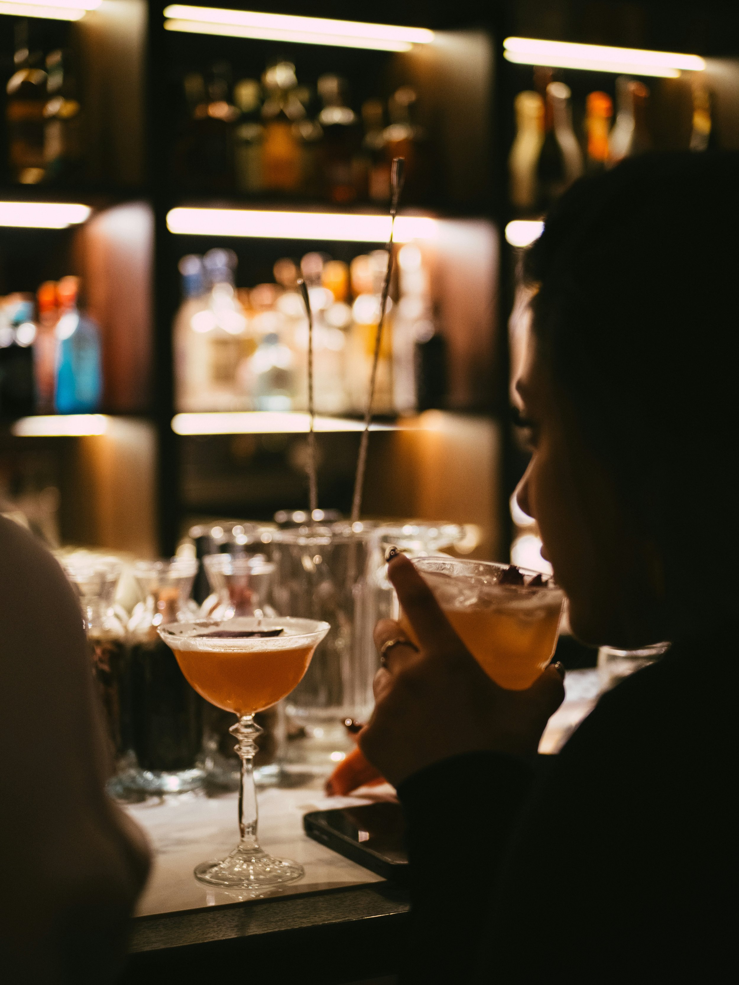 A woman drinks a cocktail at a bar with shelves of liquor bottles in the background.