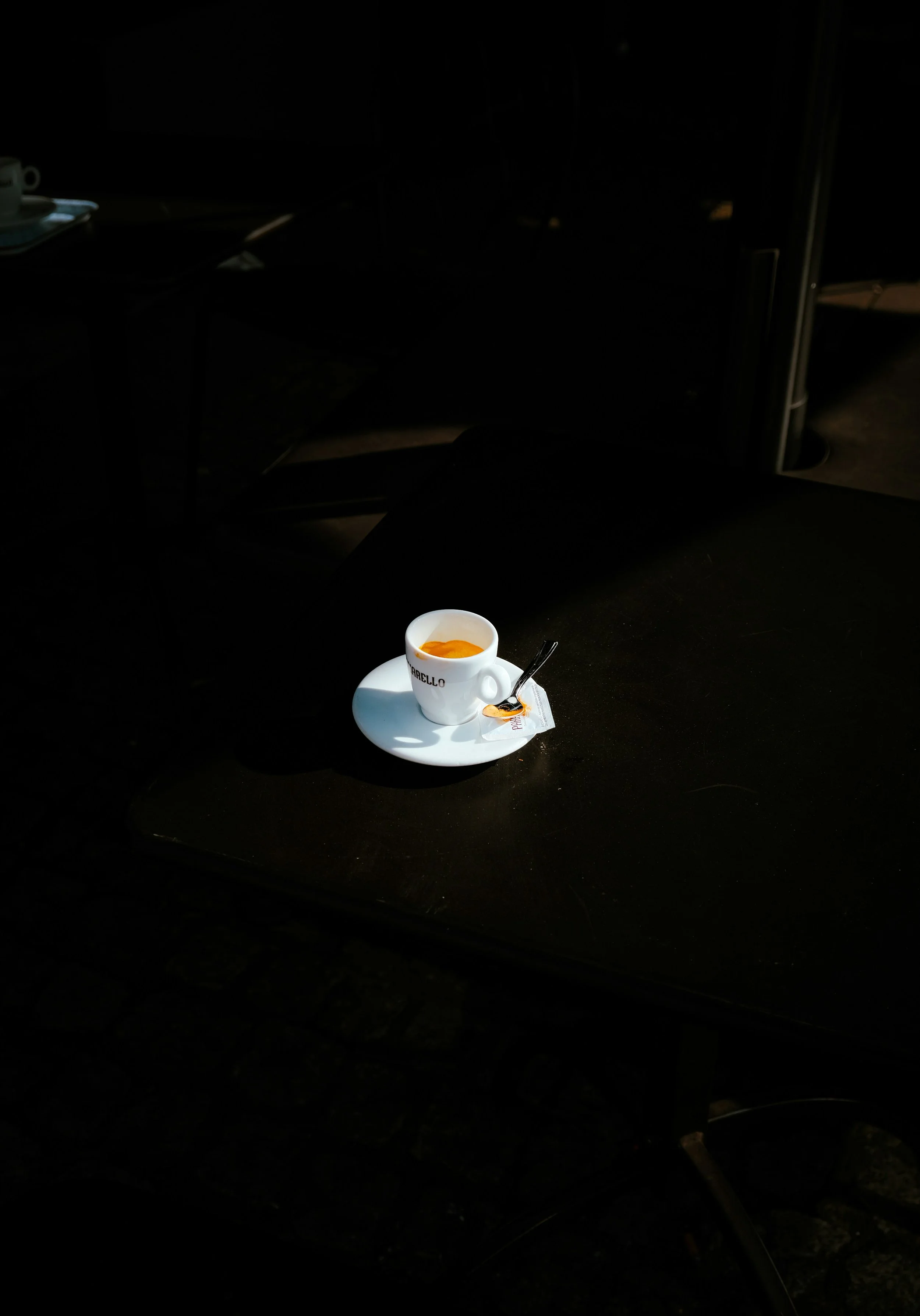 A white cup of coffee or espresso on a saucer with a small spoon and some sugar packets, sitting on a dark table in a dimly lit setting.