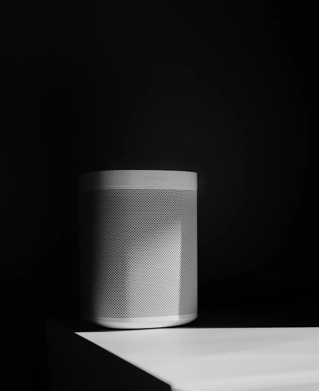 A white Sonos speaker placed on a black surface against a black background, partially illuminated by a light source.