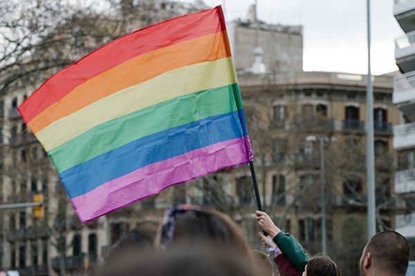 Person holding a rainbow pride flag at a public gathering in an urban area.