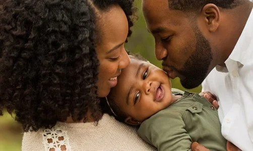 A family of three, including a woman, a man, and a young child, sharing a tender moment outdoors.