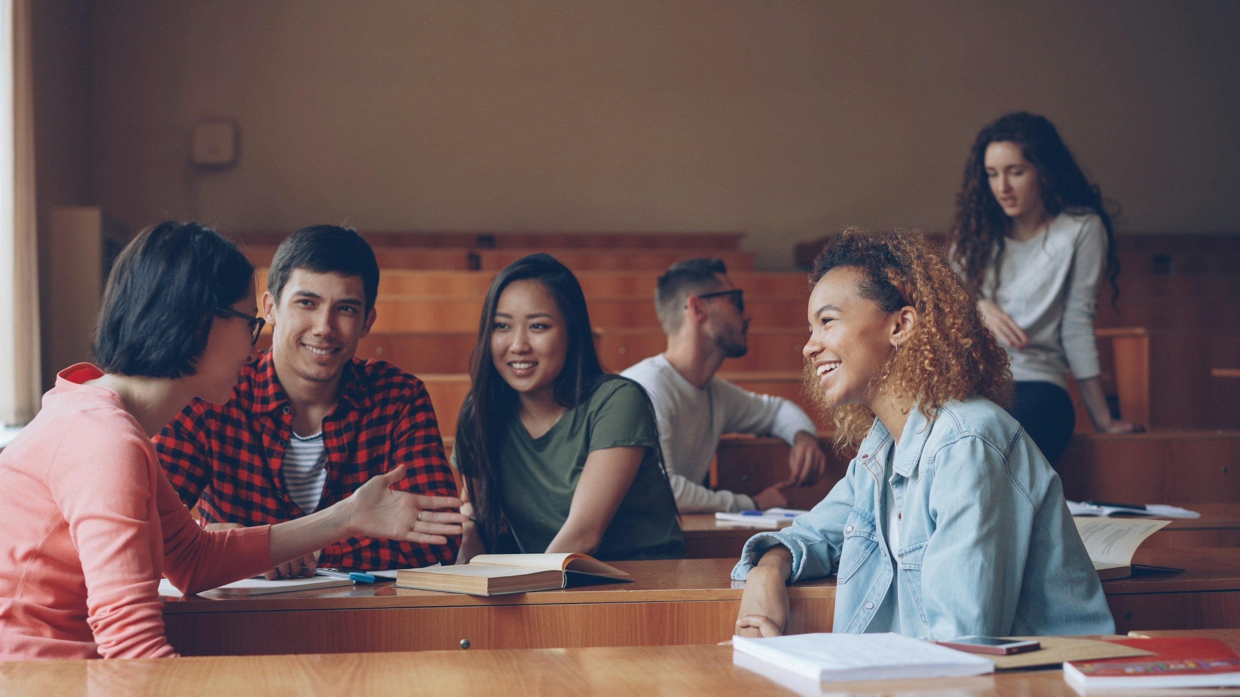 A group of diverse college students having a discussion in a lecture hall, sitting at wooden desks with open books and notebooks.