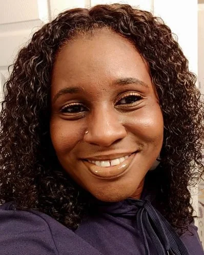 Close-up of a smiling woman with curly hair, wearing a dark top with a bow at the collar, indoors.