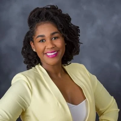 A smiling woman with curly hair wearing a light yellow blazer and a white top, posing against a gray background.