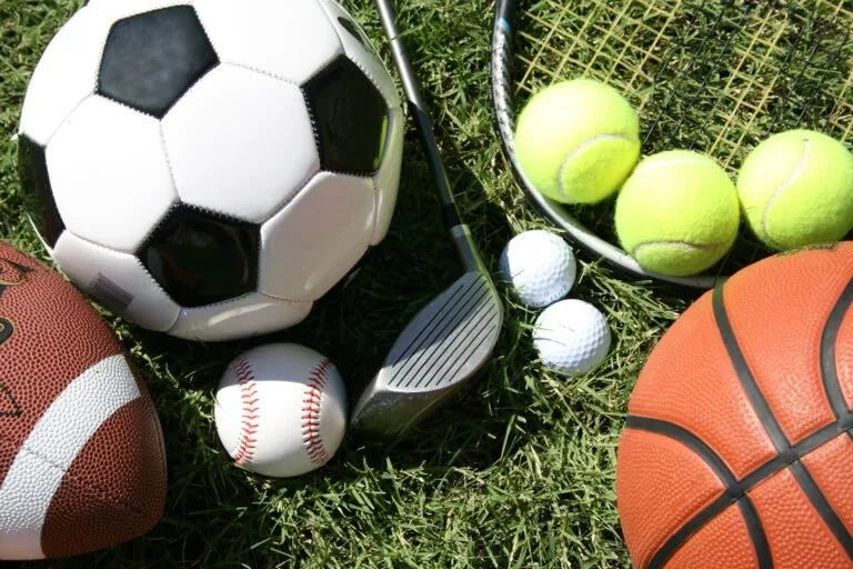 Sports equipment including a soccer ball, tennis balls, a baseball, a football, a golf club, a badminton racket, and a basketball arranged on grass.