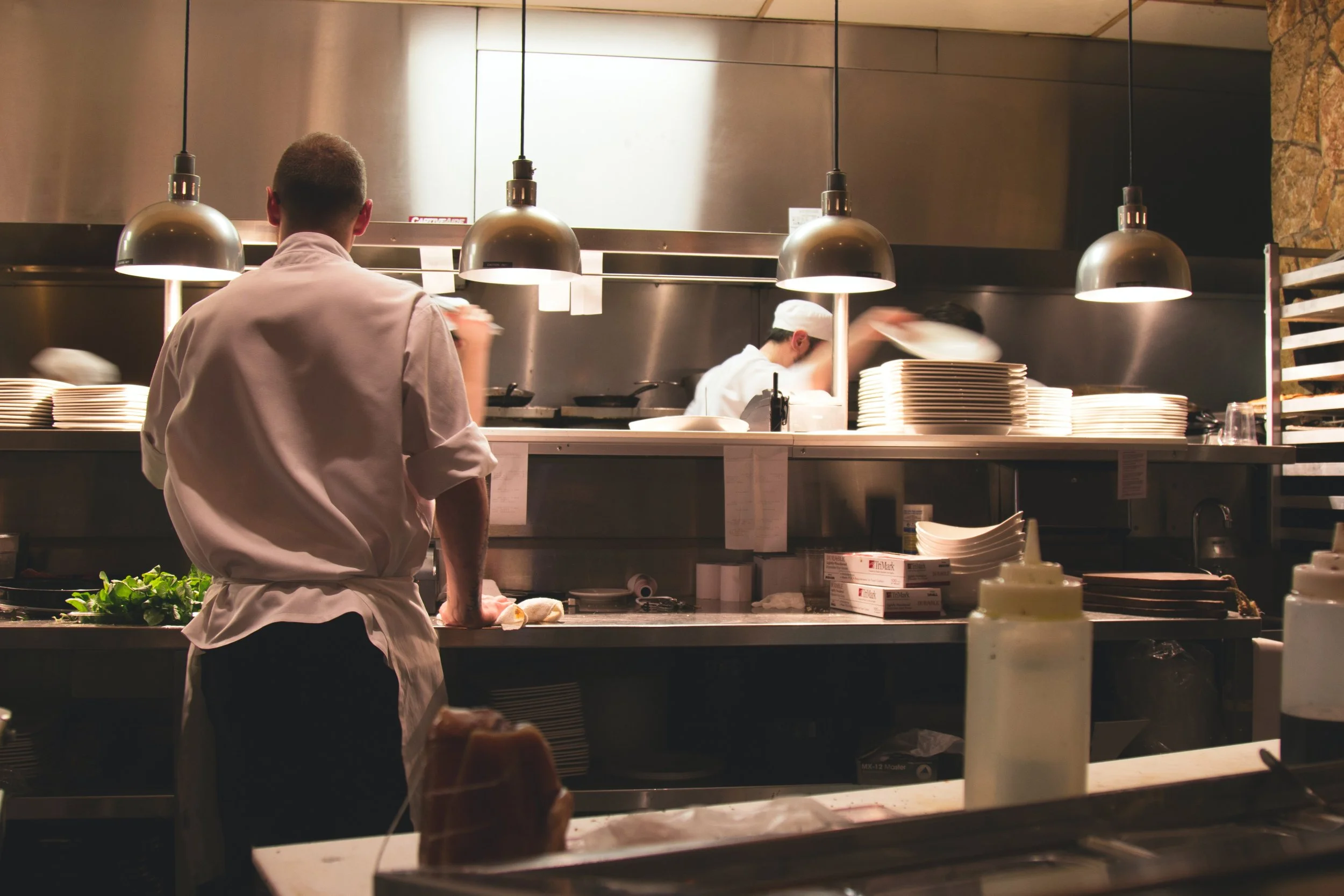 Kitchen scene with chefs preparing food under pendant lighting, plates stacked on the counter, and ingredients on the surface.