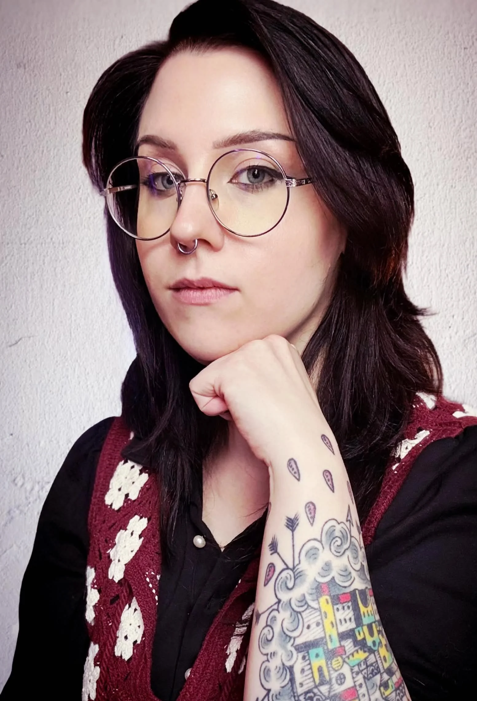 A young woman with dark hair and light gray eyes, wearing round glasses and a septum piercing, is resting her chin on her hand. She has tattoos on her arm and is dressed in a black top with a red and white patterned sleeveless cardigan. The background is a plain textured wall.