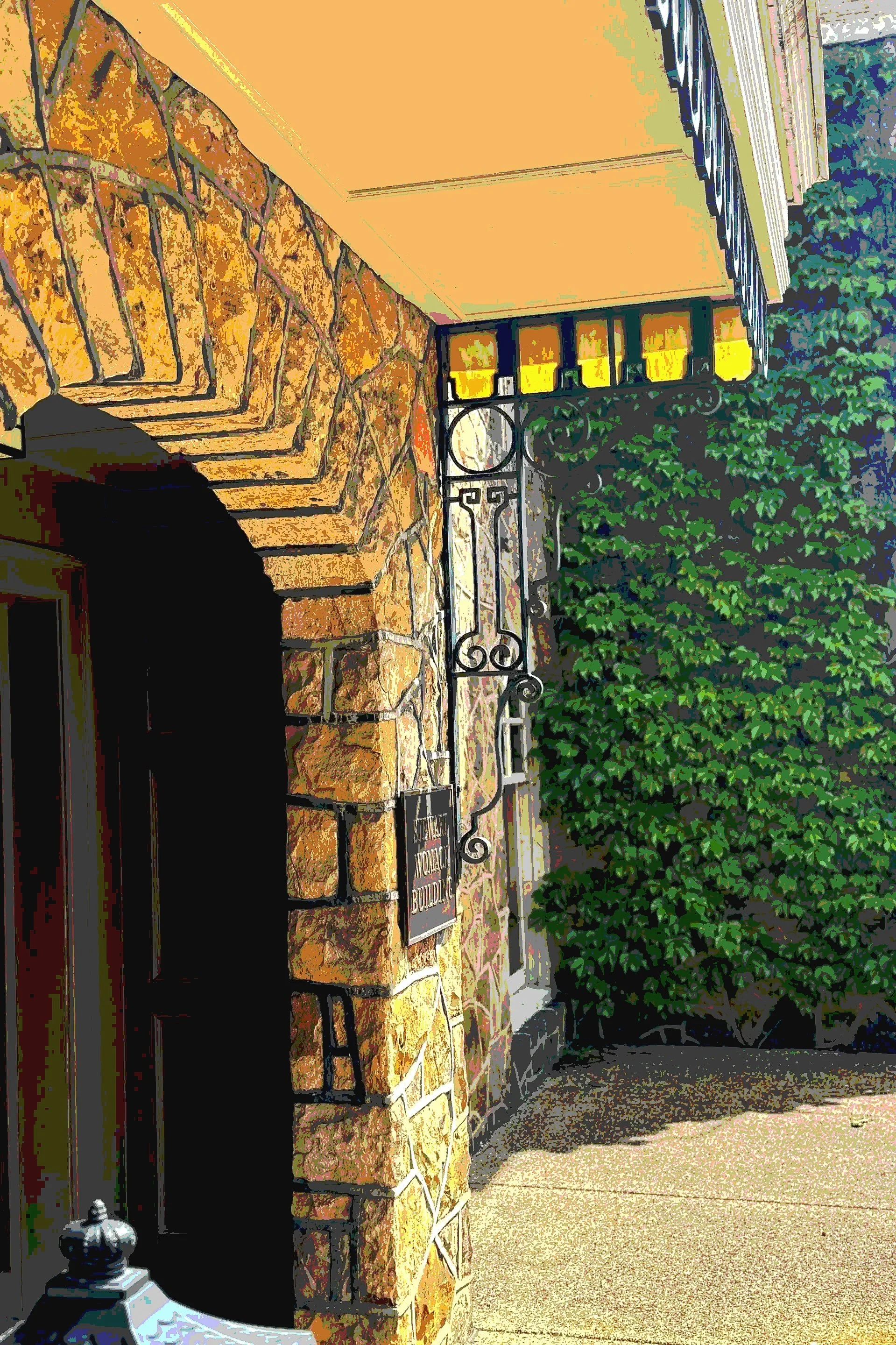 Close-up of a building corner with a stone wall, black wrought-iron gate, and green ivy on the right. The building has a beige exterior. Small sign says 'NOTICE'.
