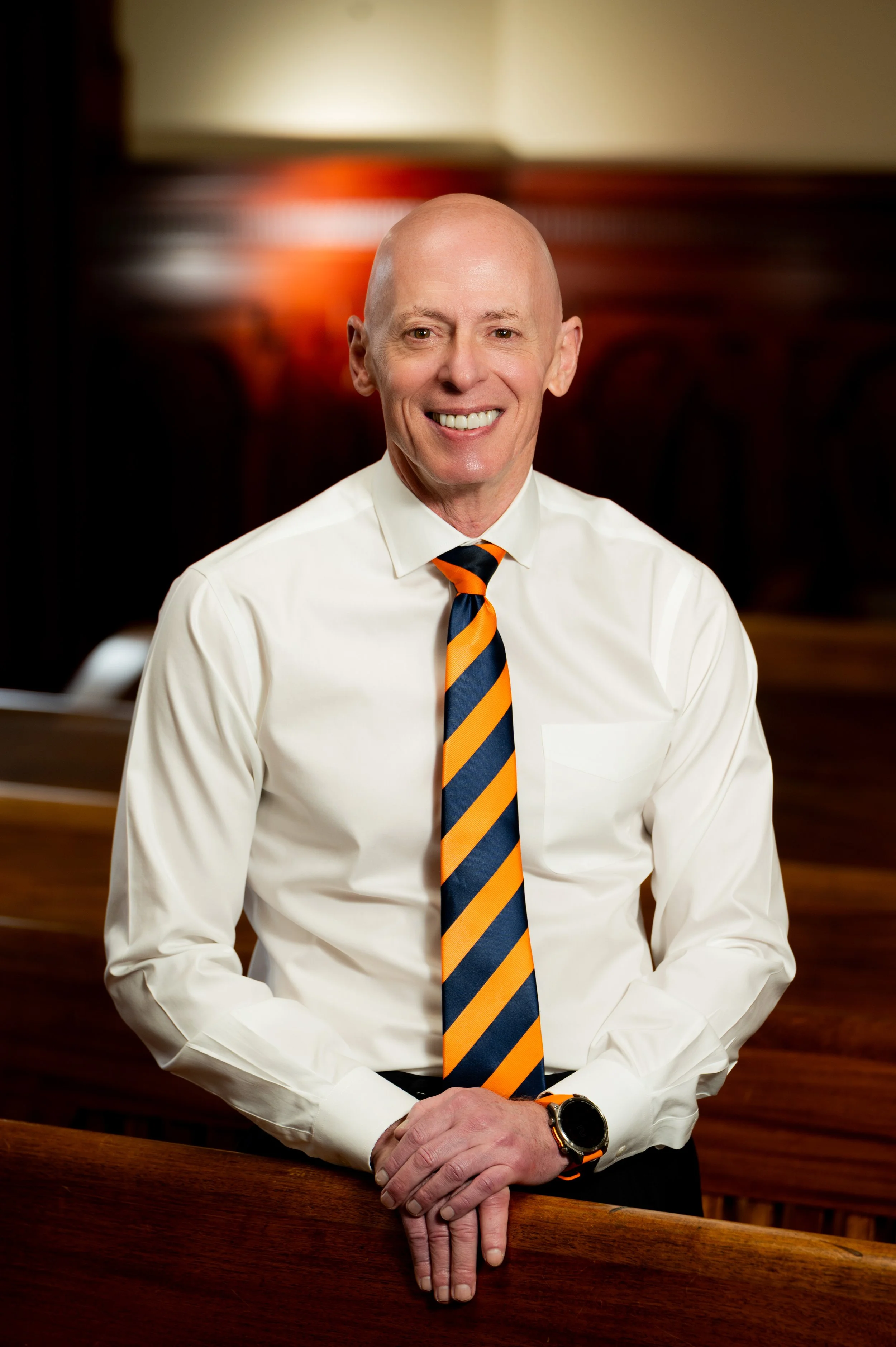A bald man in a white dress shirt and a striped orange and navy tie, smiling, with his hands folded on a wooden surface, in a warmly lit room.