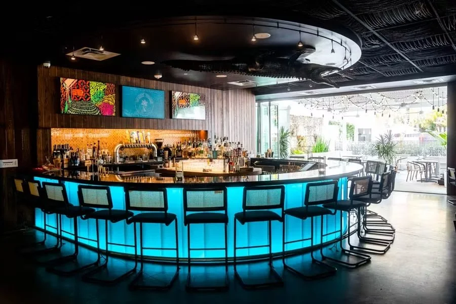 Modern bar with illuminated blue counter, bar stools, a variety of bottles behind the bar, and large windows with outdoor seating.