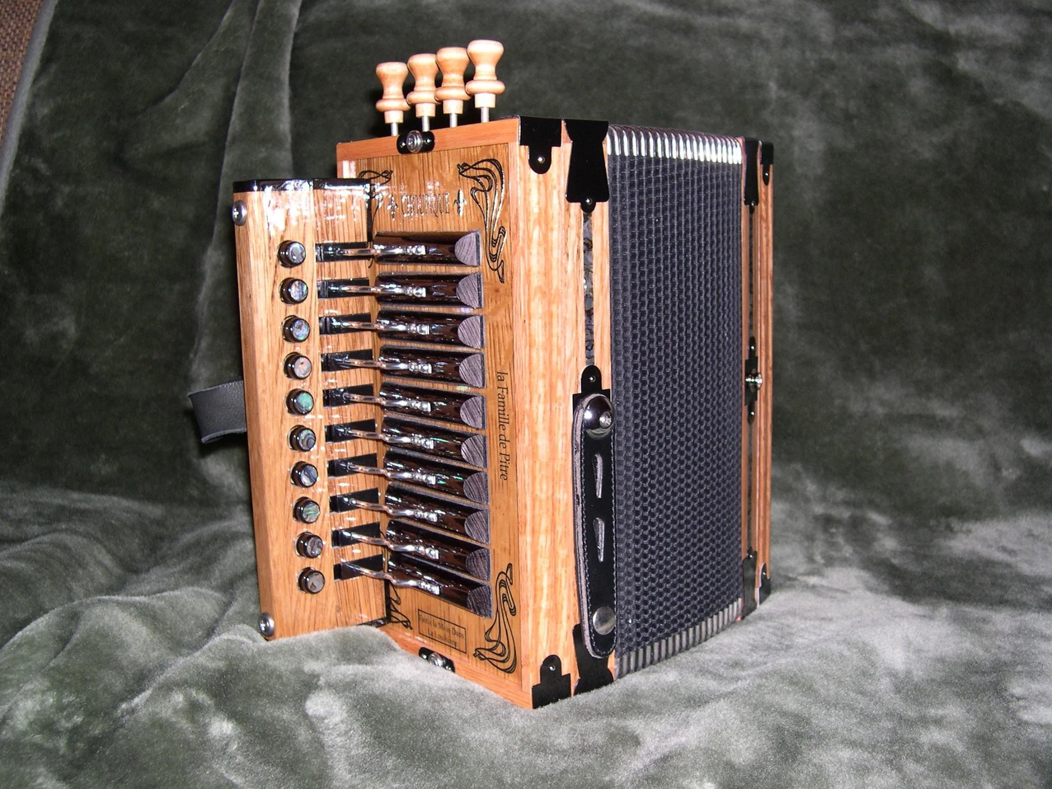 A wooden musical instrument called a Choupique Accordion with metal keys and a black reed valves, resting on a dark green velvet surface.