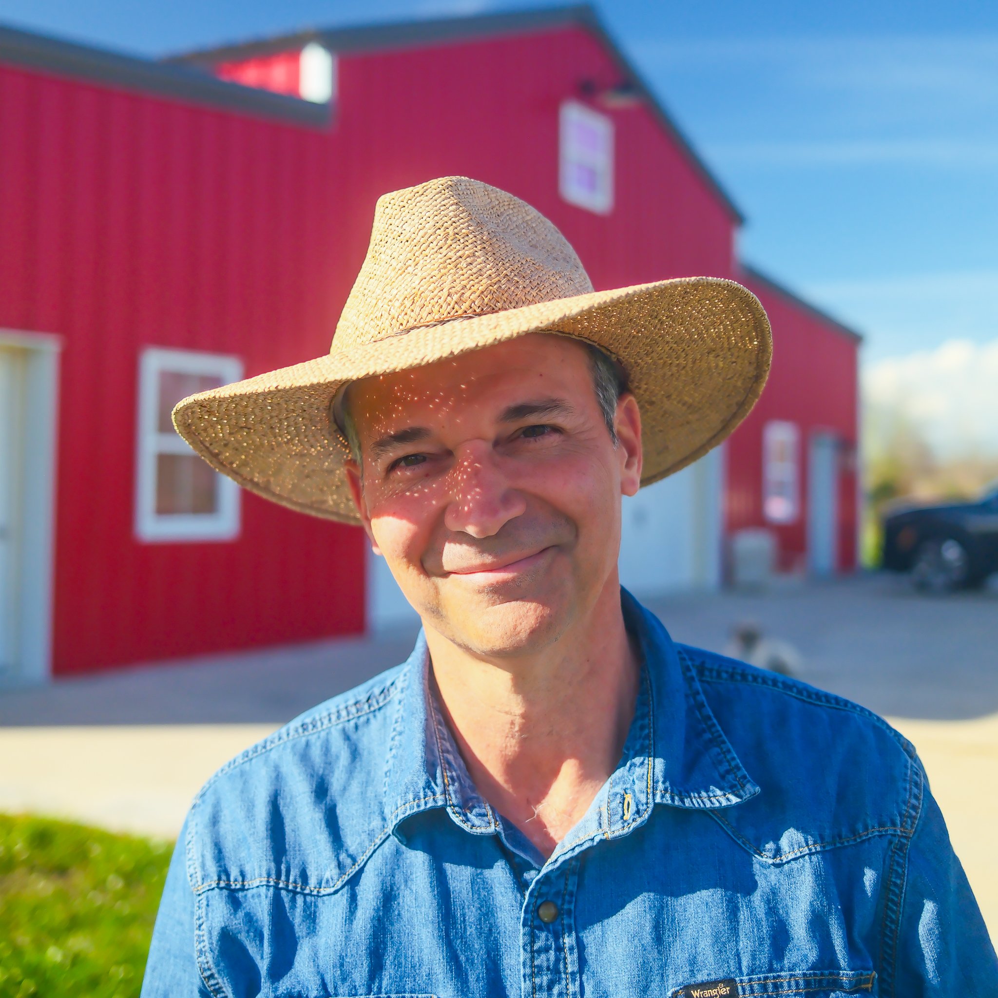 Close-up of Jesse Brown, a smiling man wearing a straw hat and a denim jacket in front of a red building on a sunny day. Choupique Caban