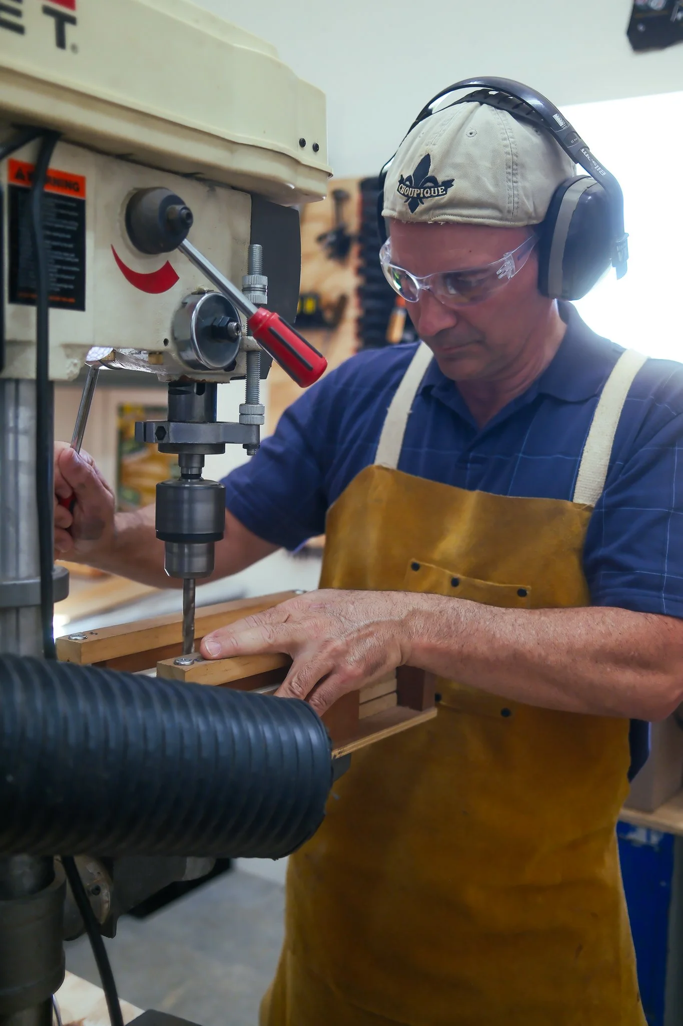 Jesse Brown is a man wearing safety glasses, headphones, and a baseball cap working with a drill press in a workshop.