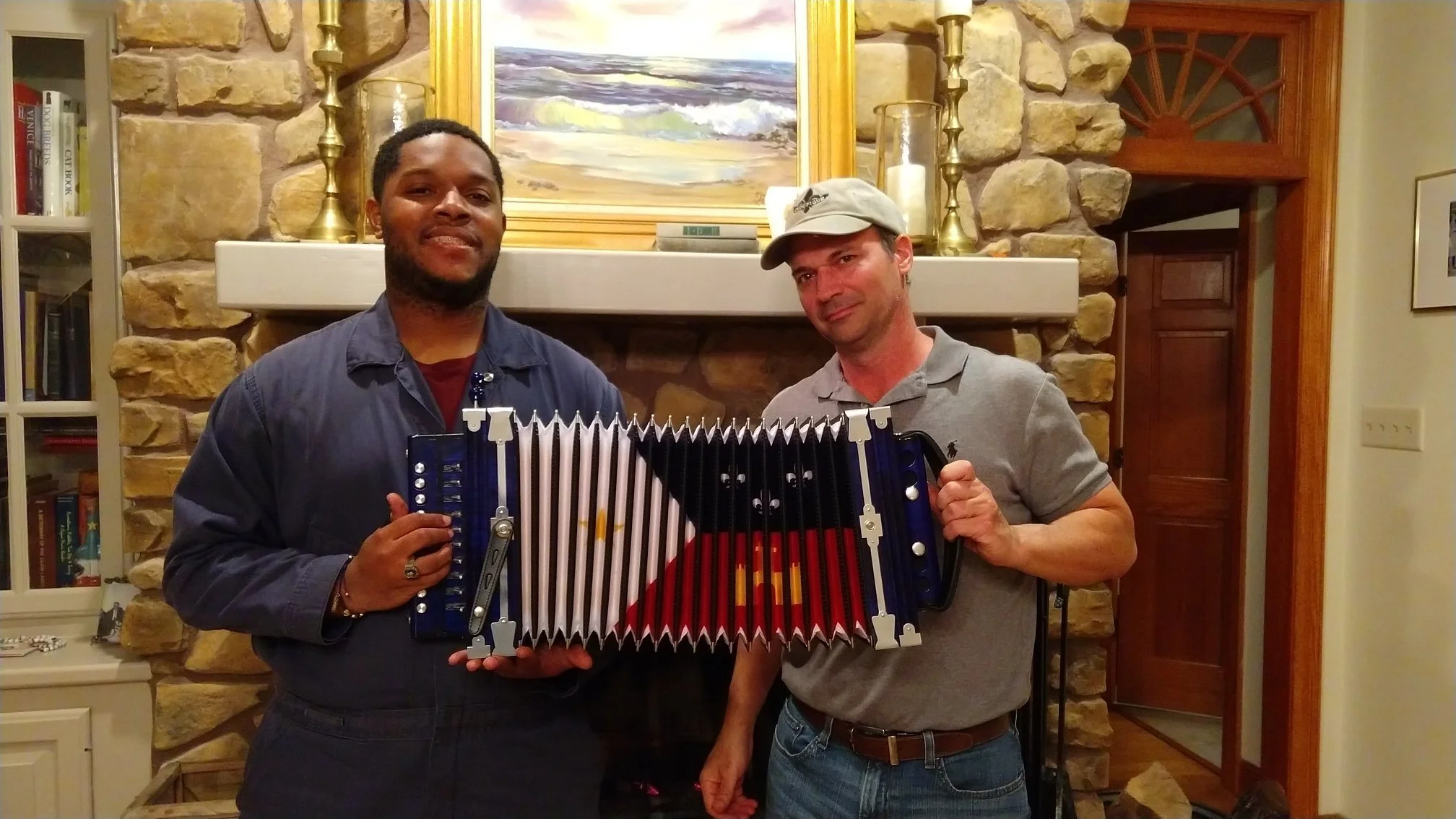 Two men standing in front of a stone fireplace holding a large accordion. The man on the left has dark hair and is wearing a navy jacket, smiling. The man on the right has light hair, is wearing a light gray polo shirt and a beige cap, and is smiling. A framed painting is above the fireplace, and candles and candlesticks are on the mantel.