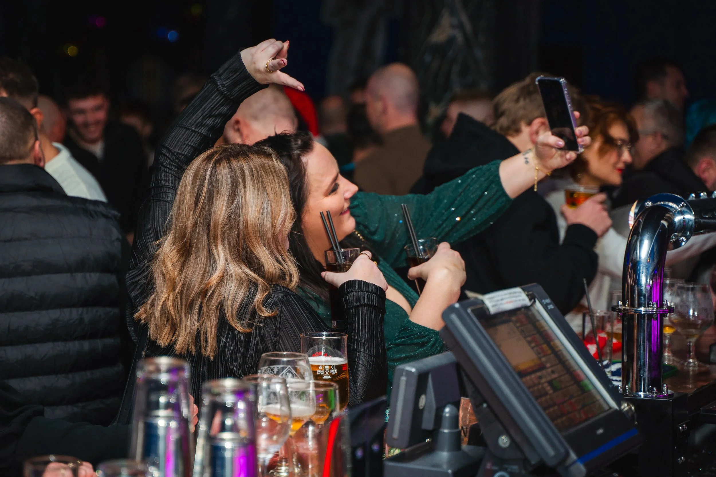 A crowded bar with people seated, some drinking and others taking photos or videos on their smartphones.