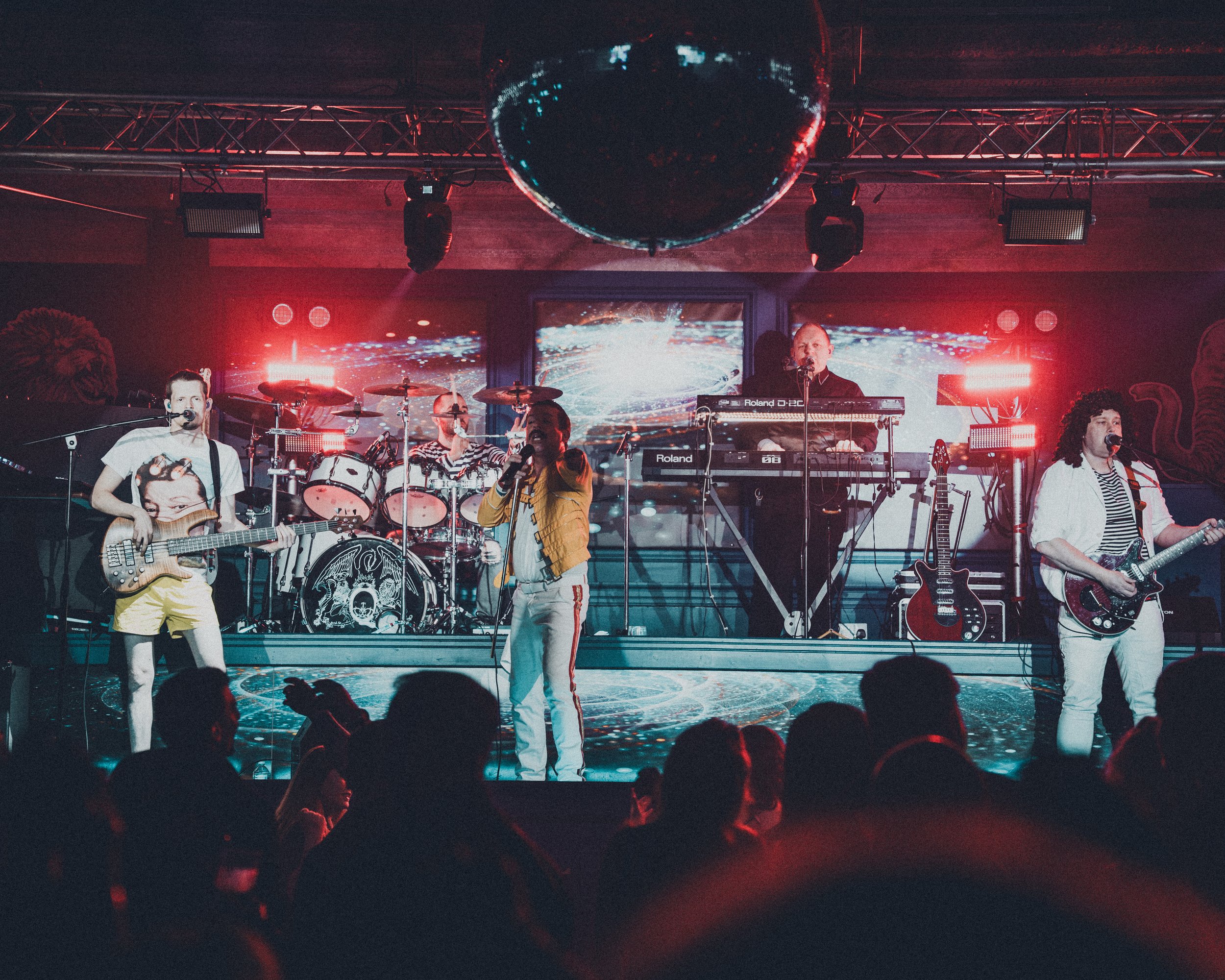 A band performing on stage with colorful lights and a large screen in the background displaying abstract visuals, and audience members in the foreground.