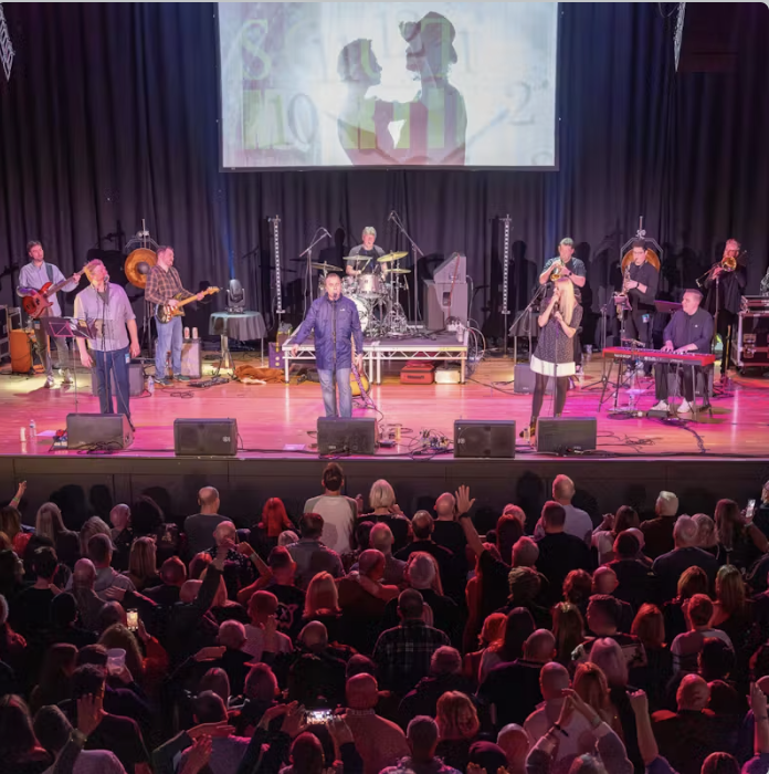 Band performing on stage with musical instruments and large audience in a theater or concert hall.