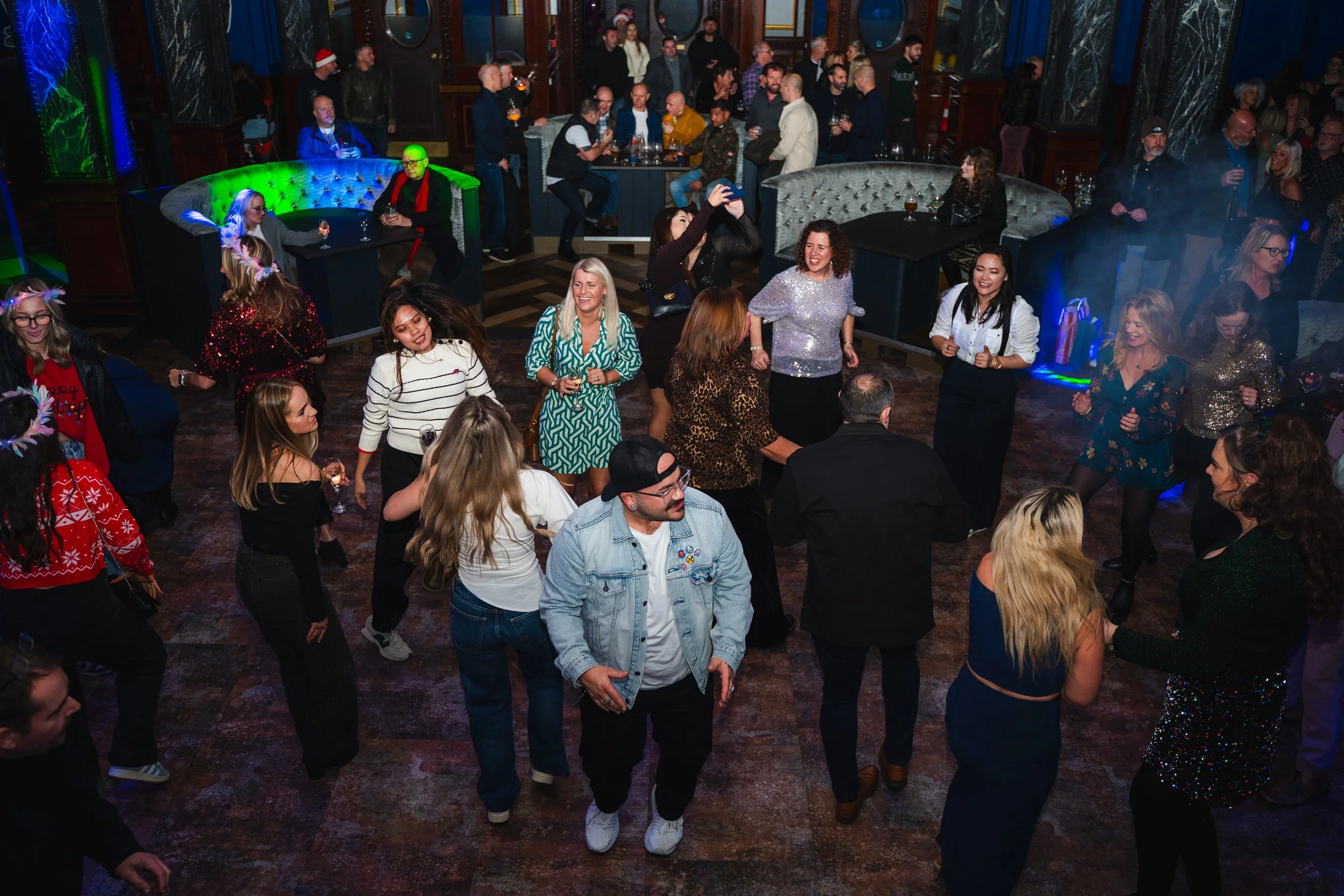 People dancing and socializing in a festive bar or club with colorful lighting, some wearing holiday-themed clothing.
