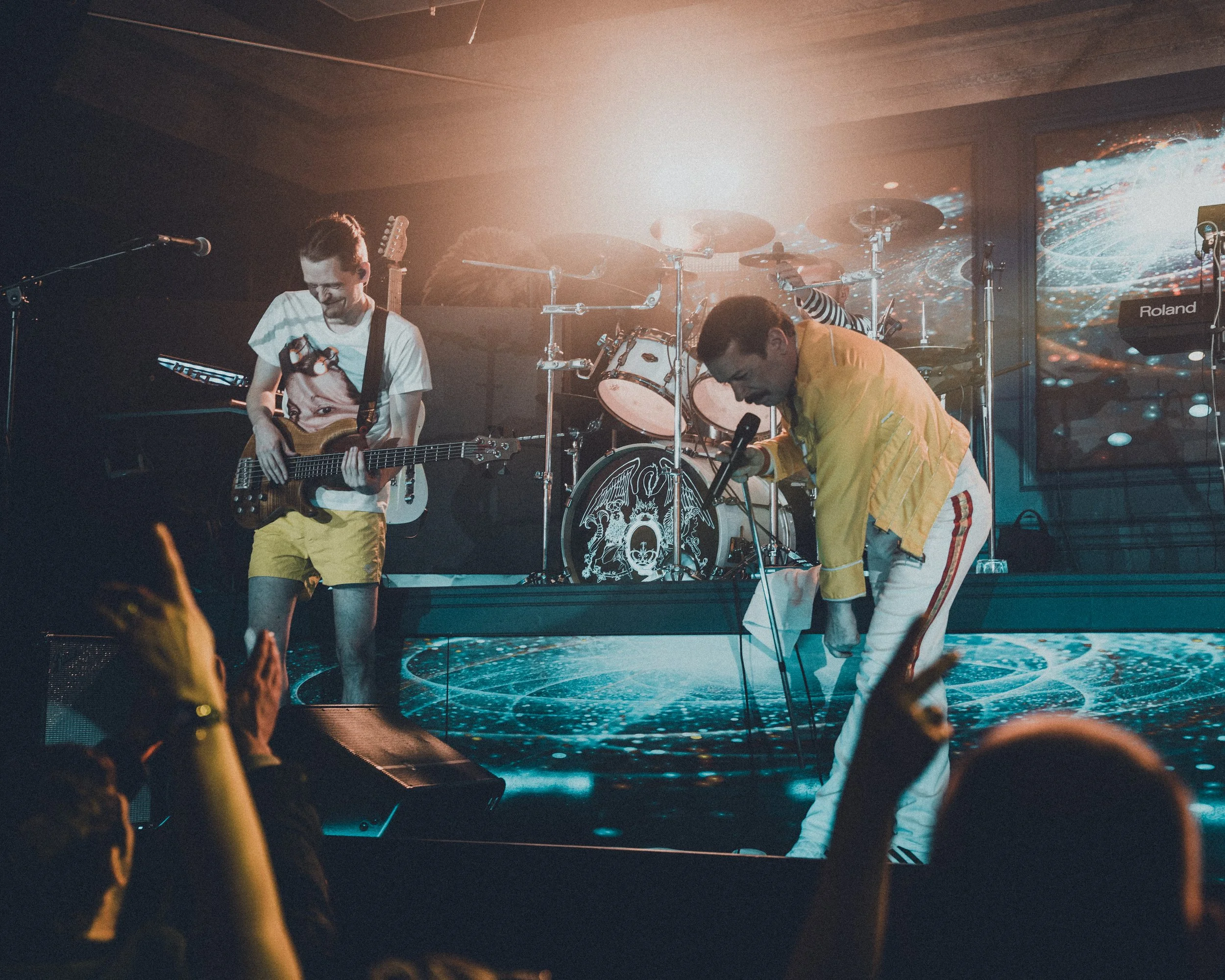 Two performers on stage during a concert, one playing guitar and the other crouching near a microphone, with drums and electronic equipment in the background and audience hands in foreground.
