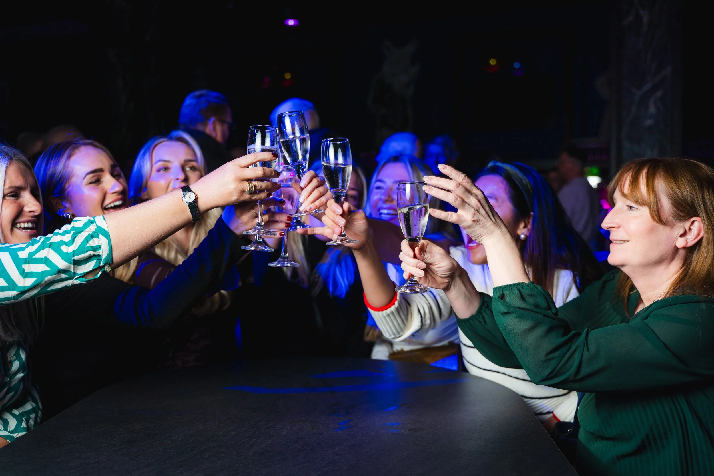 Group of women raising champagne glasses in a toast at a lively celebration or party in a darkened room with colorful lights.