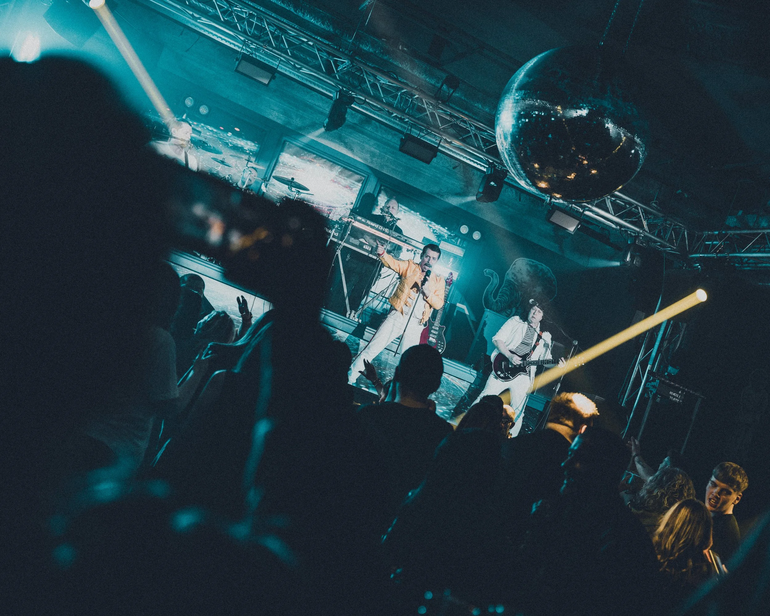 A nightclub scene with a DJ playing on a stage, a large disco ball hanging from the ceiling, and a crowd of people dancing and raising their hands in the foreground. The lighting is dim with blue and yellow highlights.