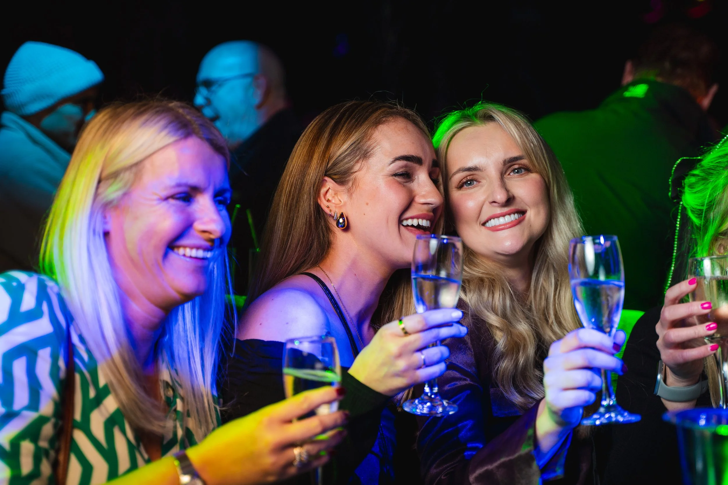 Group of women at a celebration, holding champagne glasses, smiling, with colorful lighting in a lively setting.