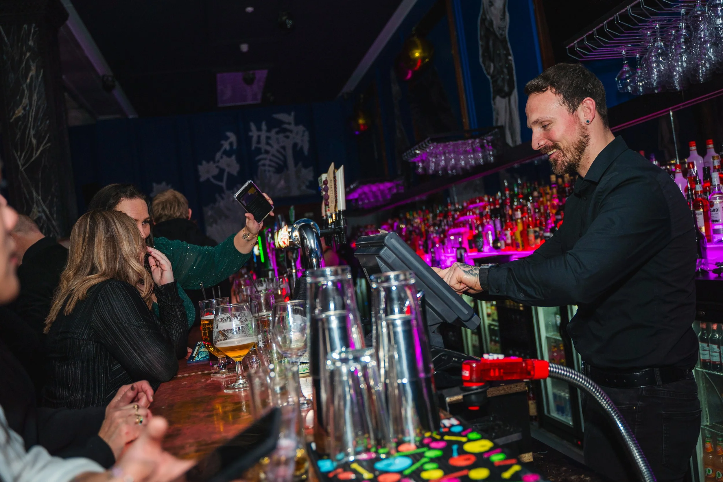 A bartender serving drinks to a group of women at a bar with colorful neon lighting.