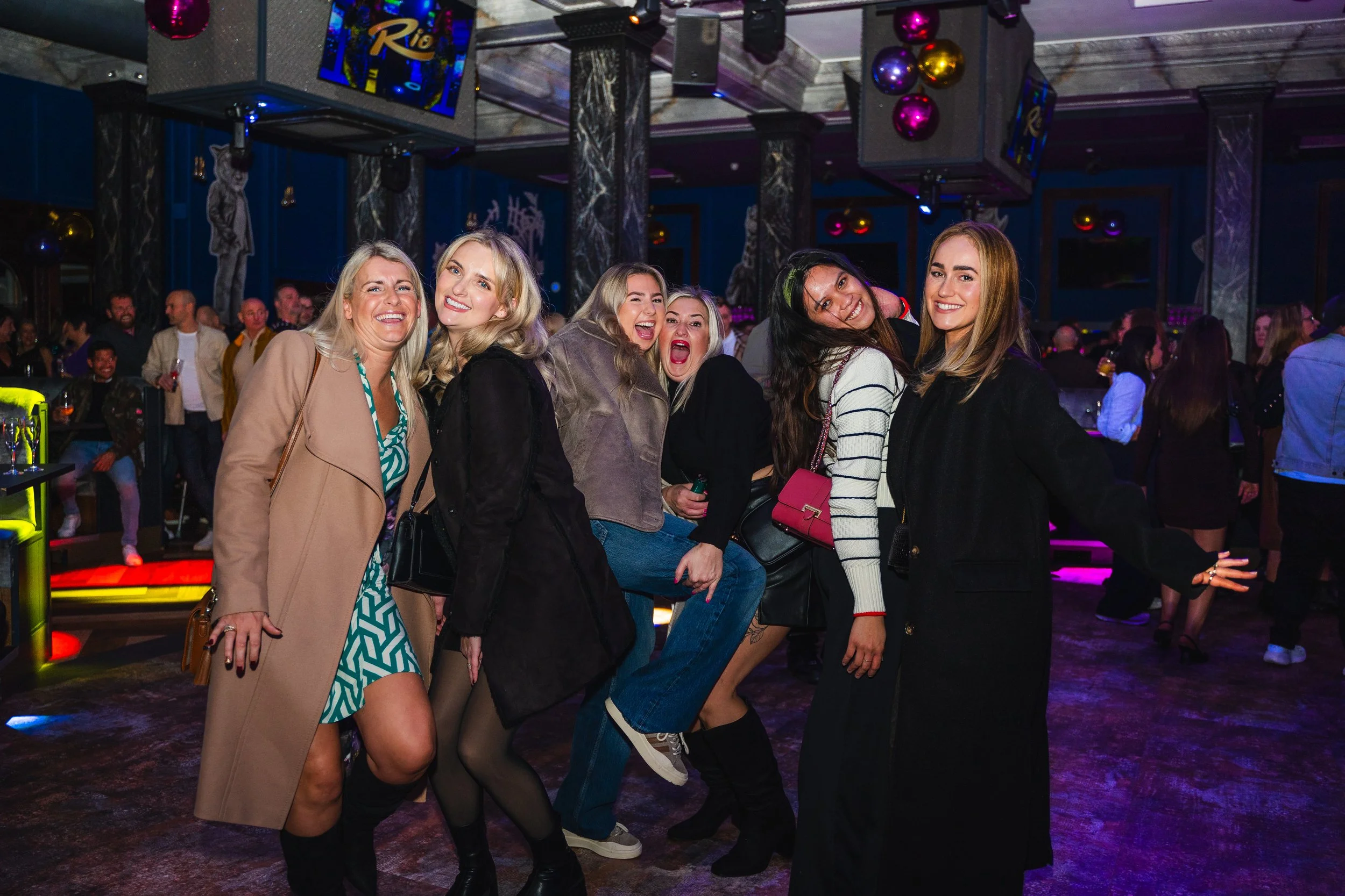 Group of seven women smiling and posing at a party or nightclub with colorful lighting and a crowded background.