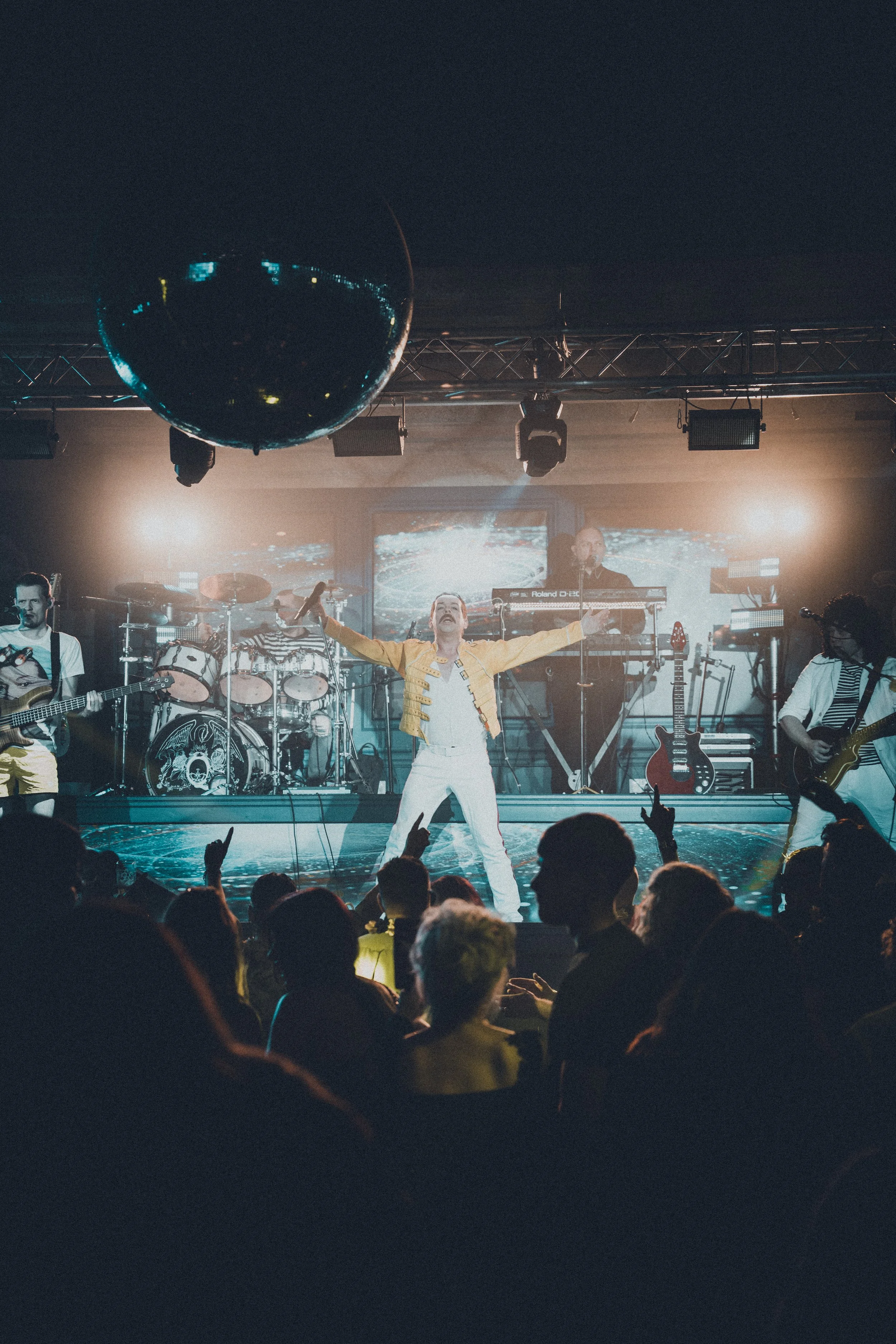 A concert stage with a singer in a yellow jacket and white pants, arms outstretched, with a band playing behind. Audience members are visible in the foreground with raised hands, and stage lighting creates a lively atmosphere.