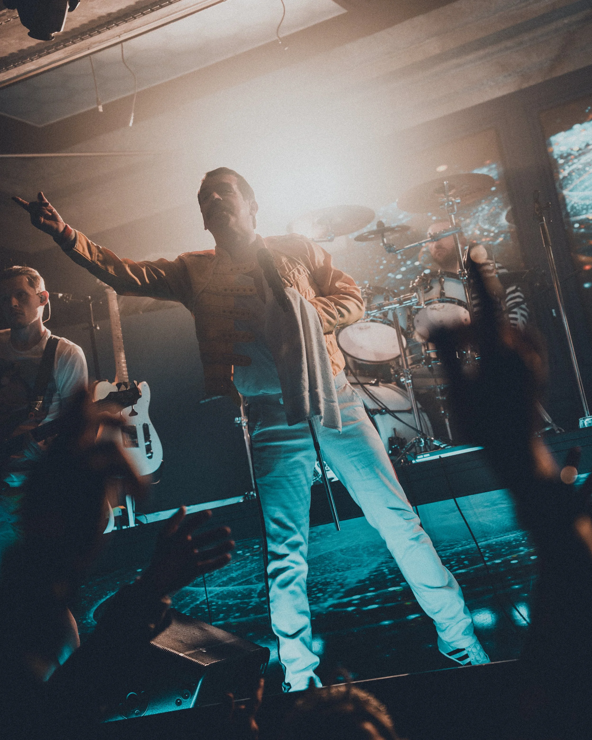 A person in a yellow racing suit with small ash-colored stripe details on the side, a gray towel draped over one shoulder, and white pants, standing on stage with a microphone, pointing to the audience. Behind them, a drummer plays drums, and a perso