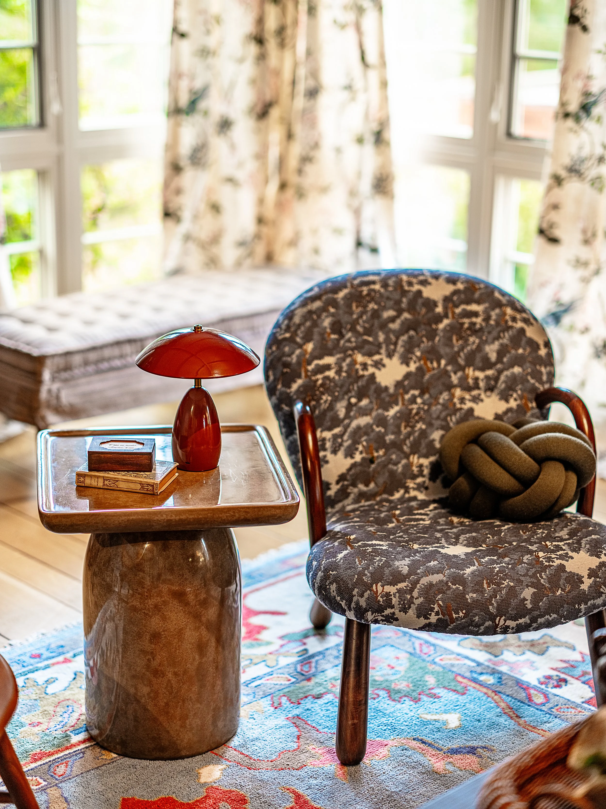 A cozy corner of a room with a floral patterned armchair and large windows with floral curtains allowing natural light. Hannah Lowe Interiors is a luxury interior designer in San Francisco and beyond.