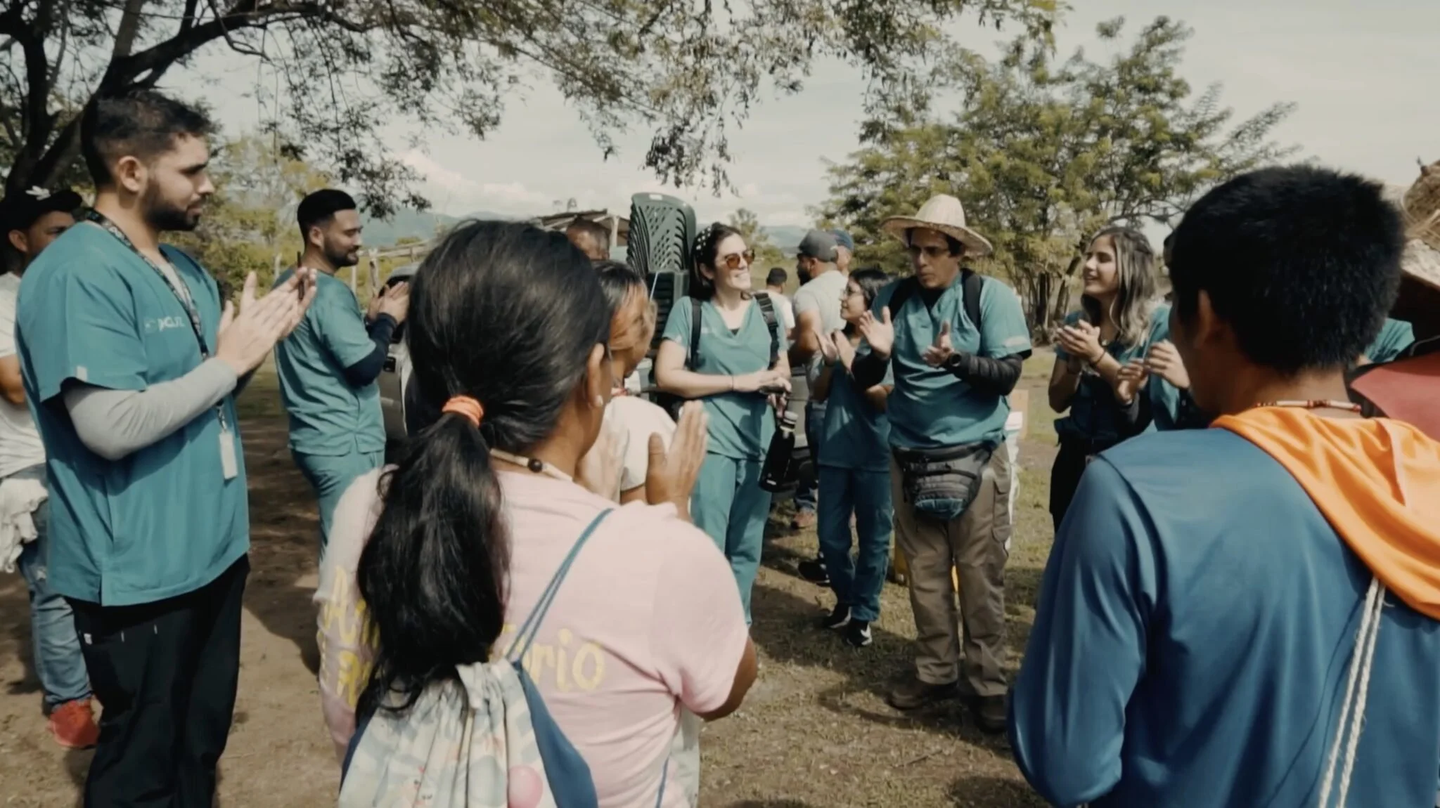 A group of people standing outdoors in a semi-circle, engaged in conversation on a sunny day with trees and mountains in the background.