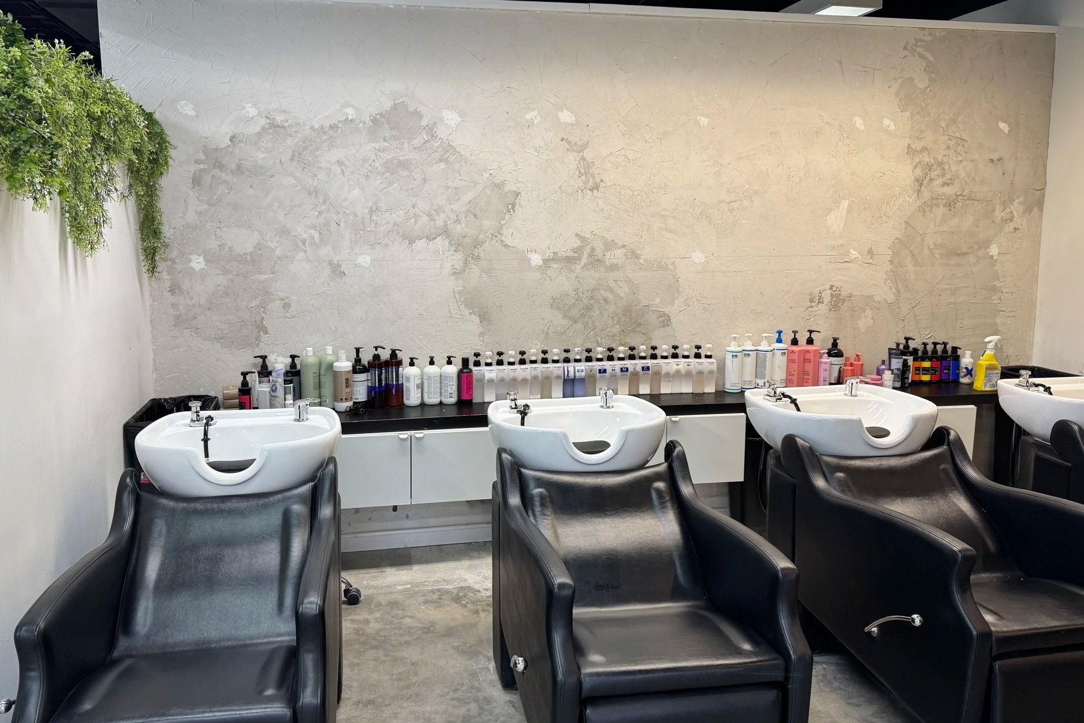 Hair salon washing station with black chairs and white sinks, shelves with hair care products on back wall.