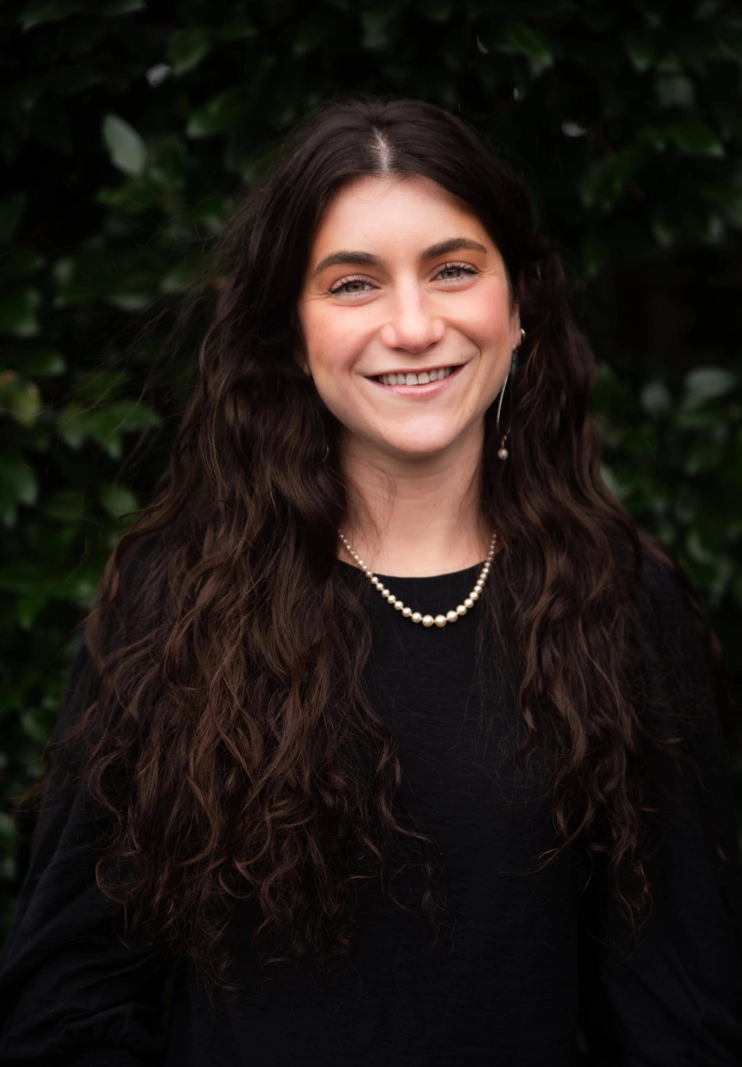 A young woman with long, wavy dark hair wearing a black top, pearl necklace, and earrings, smiling in front of green foliage.