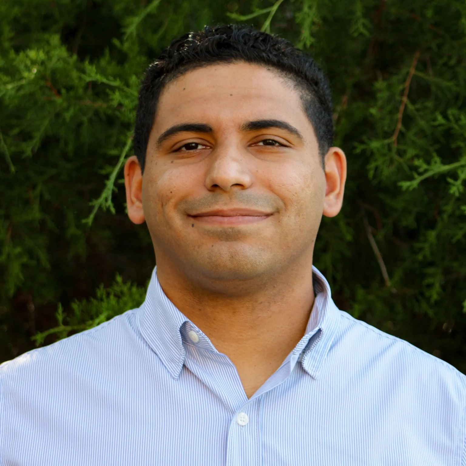 A man with short dark curly hair, wearing a light blue collared shirt, smiling in front of greenery.