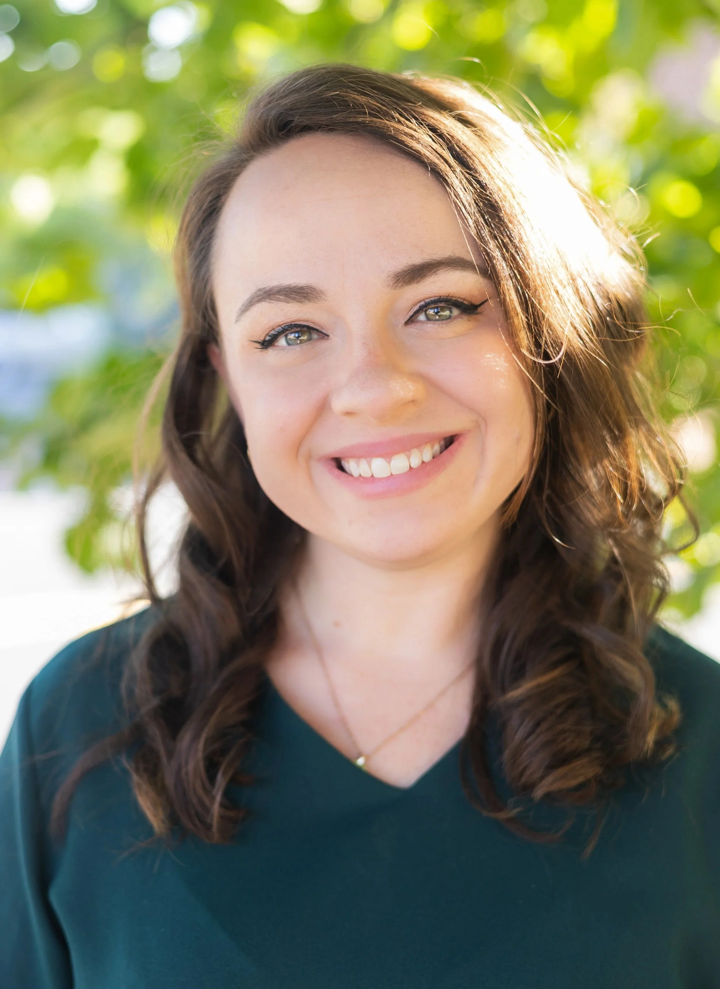A young woman with brown hair and blue eyes smiling outdoors on a sunny day with green leaves in the background.