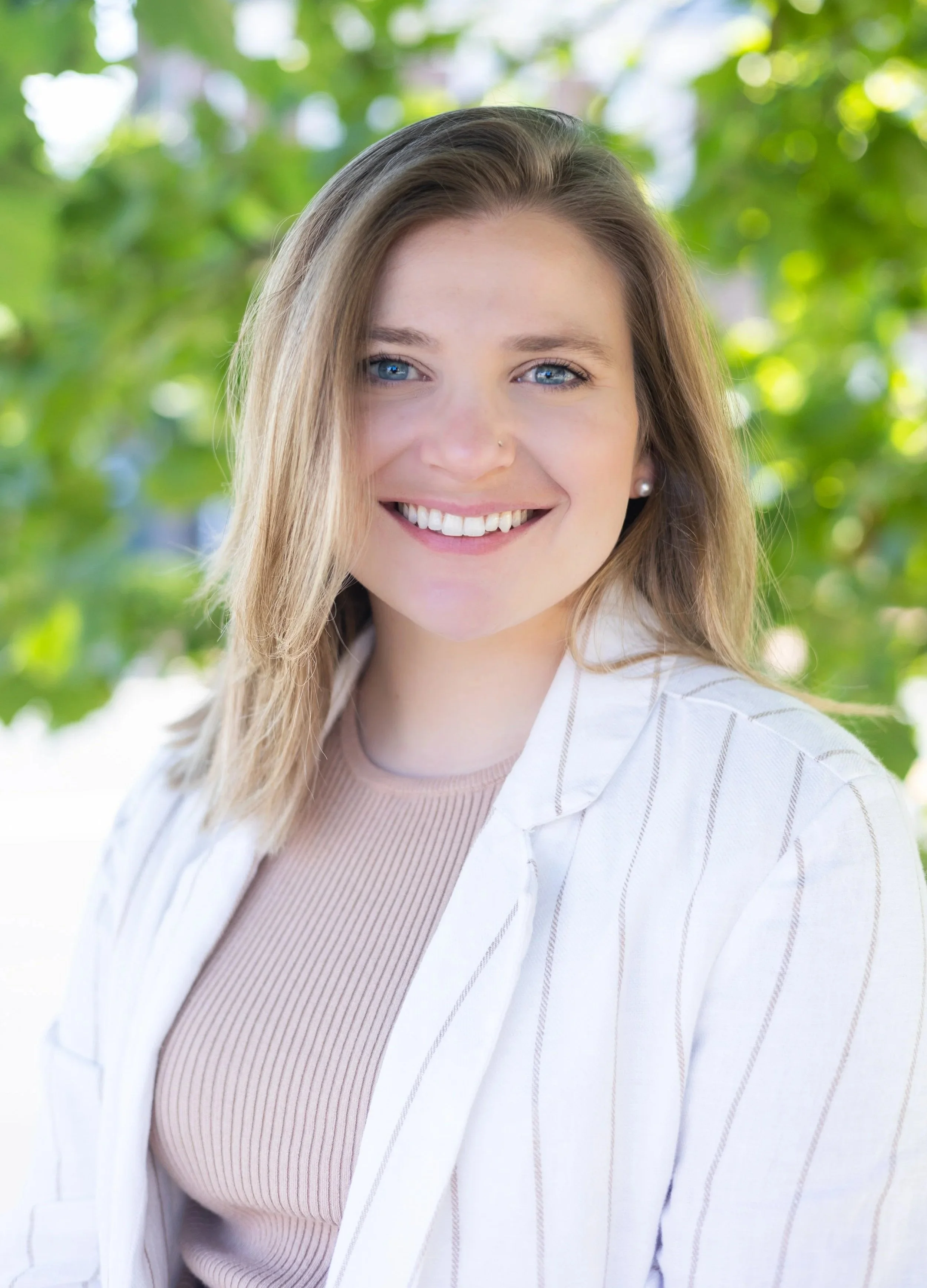 A smiling woman with blue eyes and blonde hair posing outdoors with green foliage in the background.