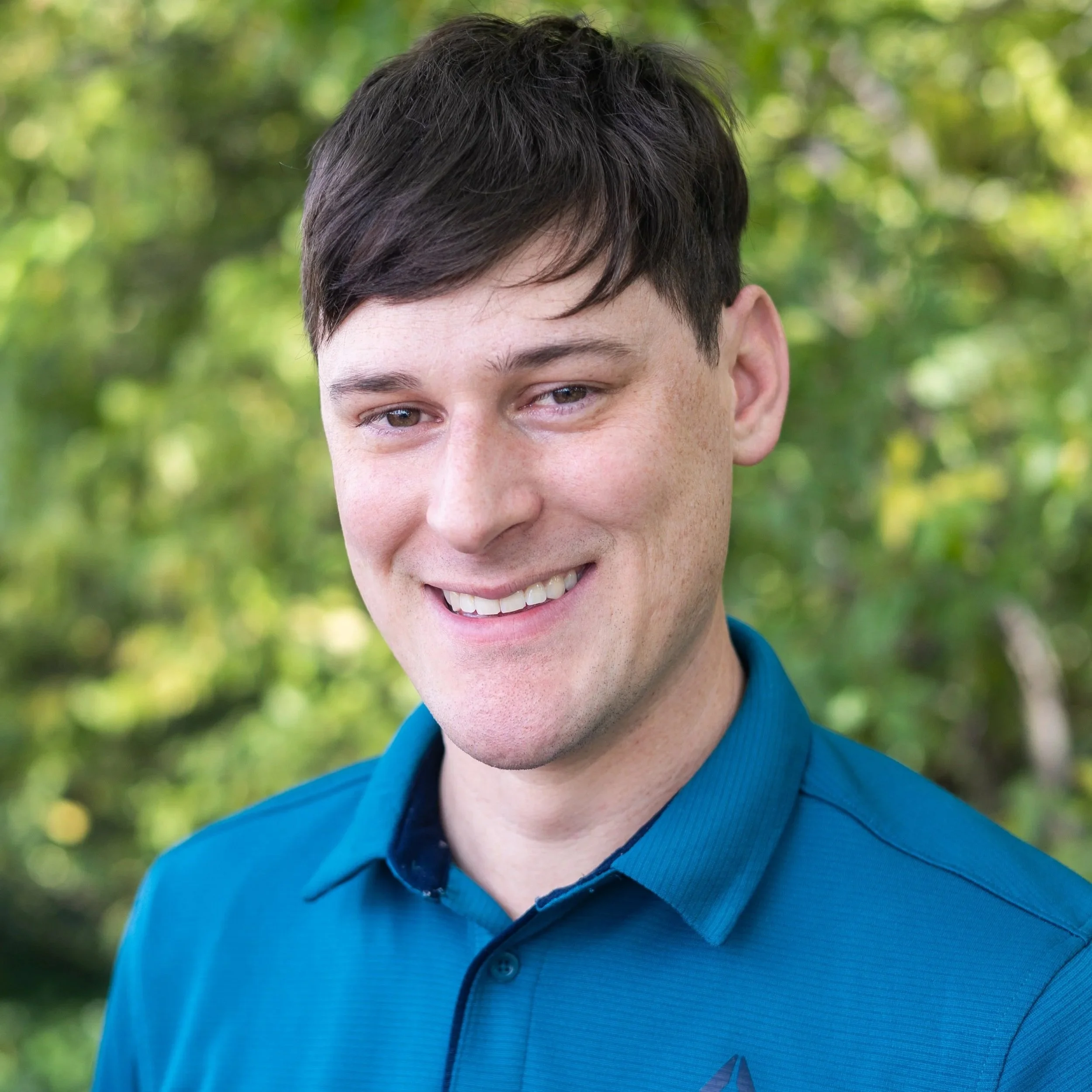 A young man with dark hair, smiling while standing outdoors with green trees in the background, wearing a blue collared shirt.