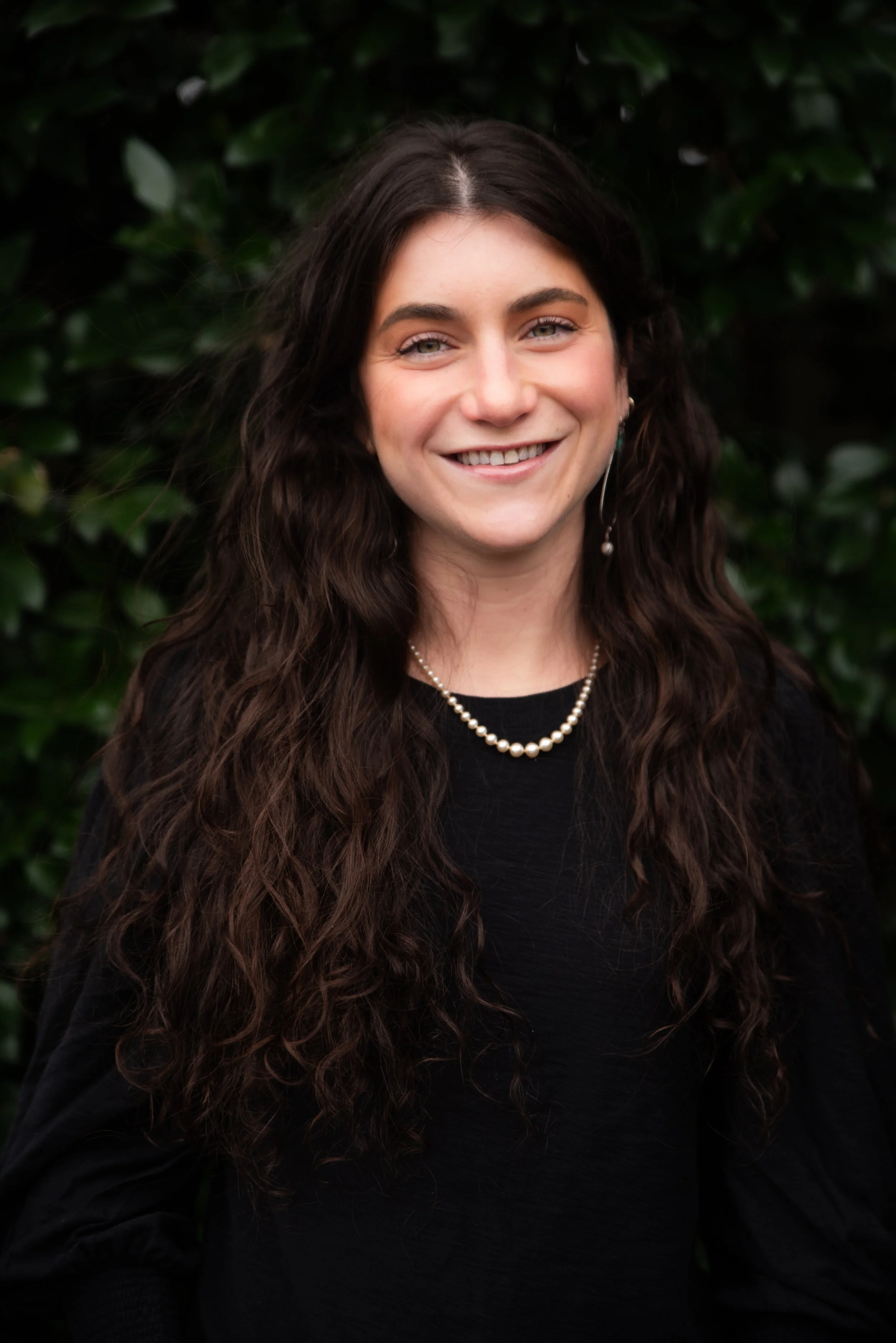 A young woman with long, curly brown hair, smiling, wearing a black top, pearl necklace, and earrings, standing outdoors in front of green foliage.