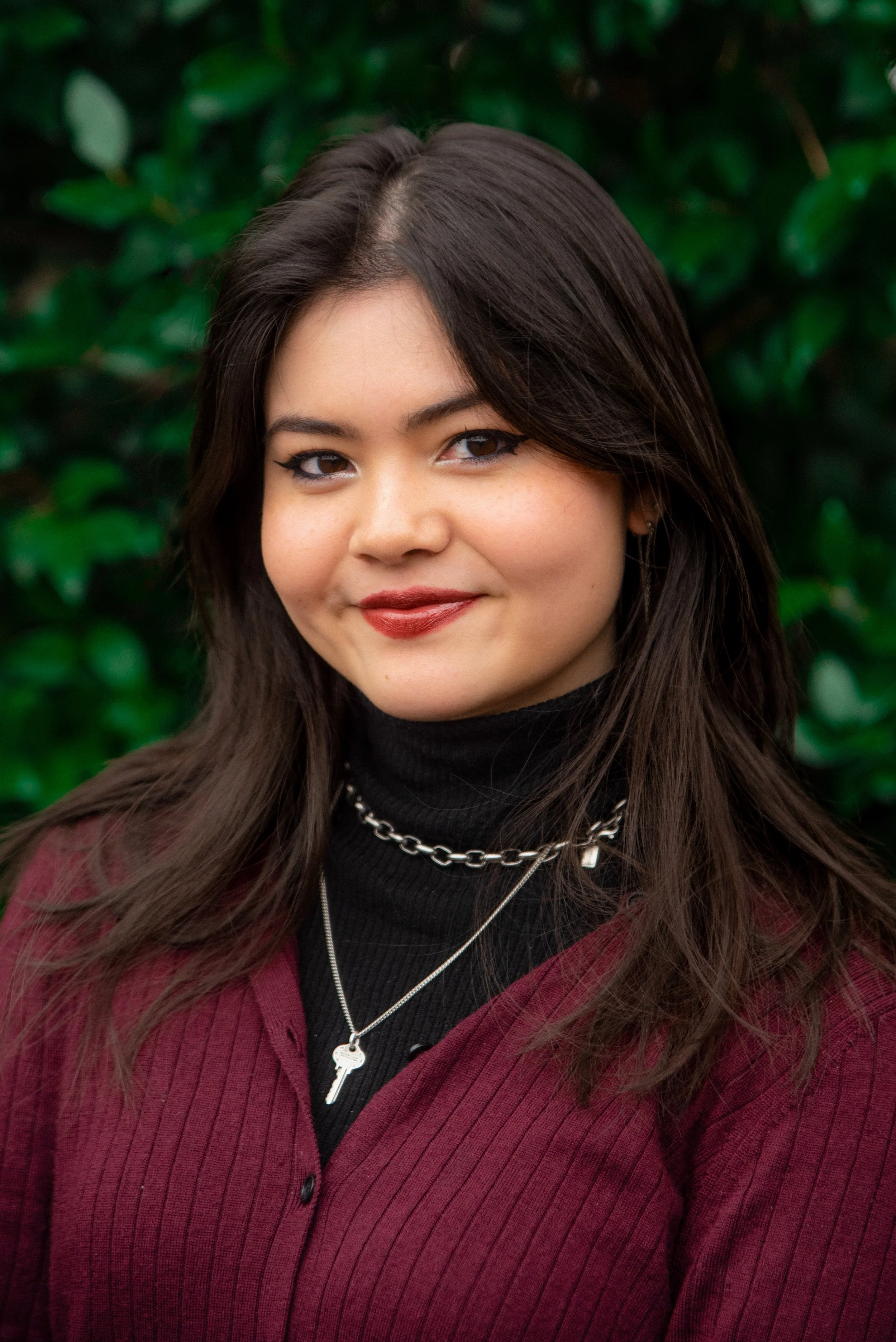 A young woman with dark hair and makeup, wearing a black turtleneck, silver necklances, and a burgundy sweater, smiling outdoors with green foliage in the background.