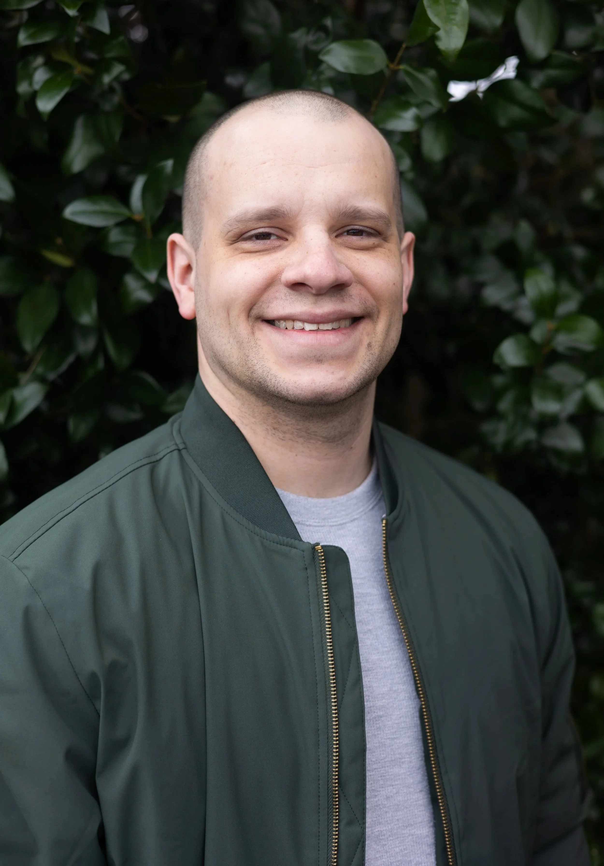 A man with a shaved head smiling, wearing a dark green jacket over a light gray shirt, standing outdoors in front of green leafy bushes.
