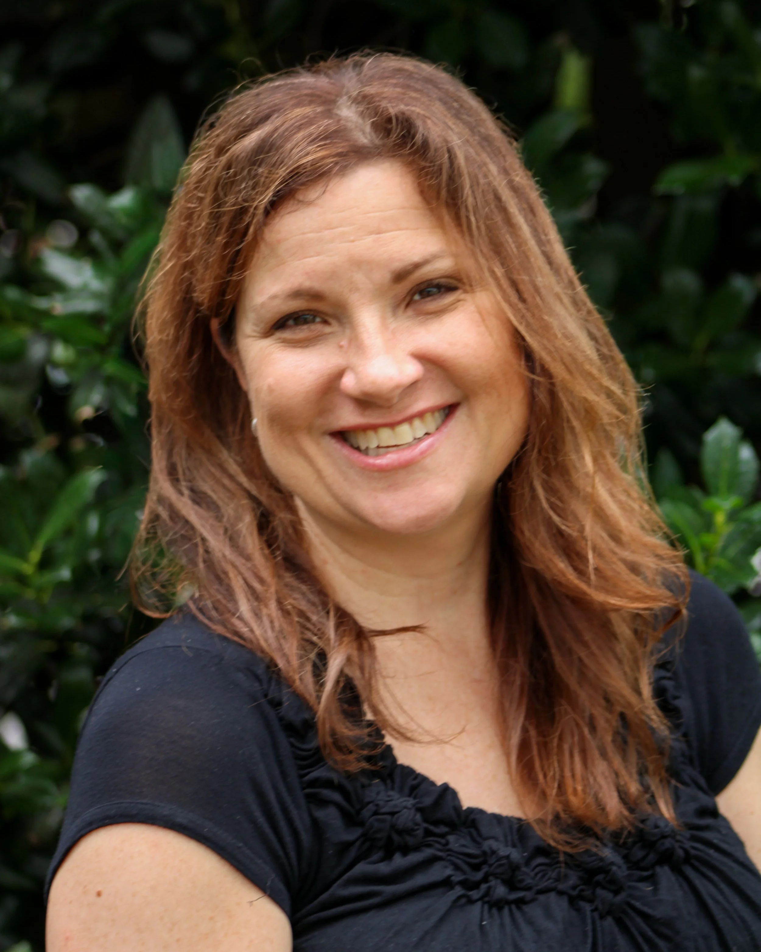 Close-up of a smiling woman with long red hair, wearing a black top, outdoors with green foliage in the background.