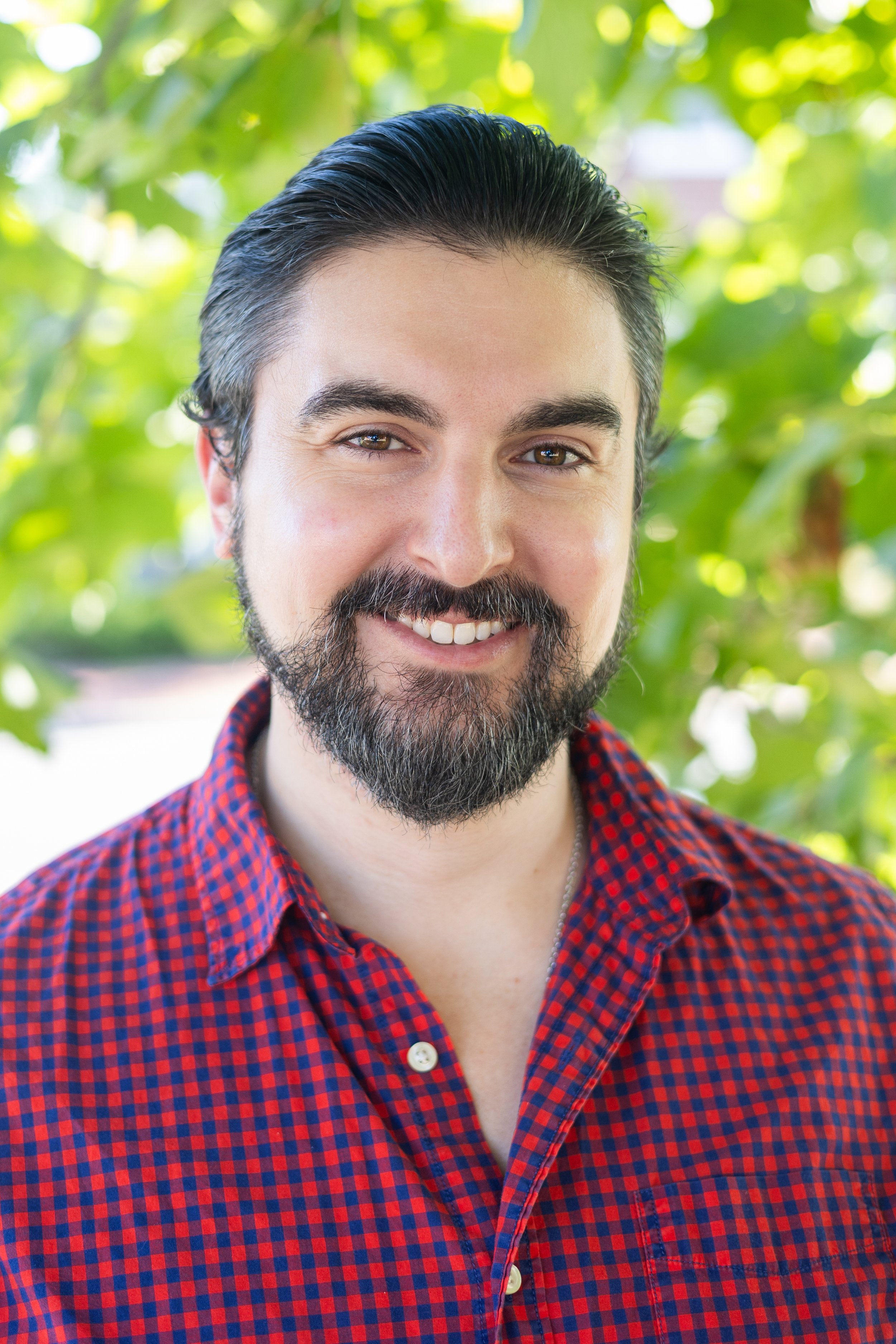 A man with dark hair, a beard, and a mustache, wearing a red and blue checkered shirt, smiling outdoors with green leaves in the background.