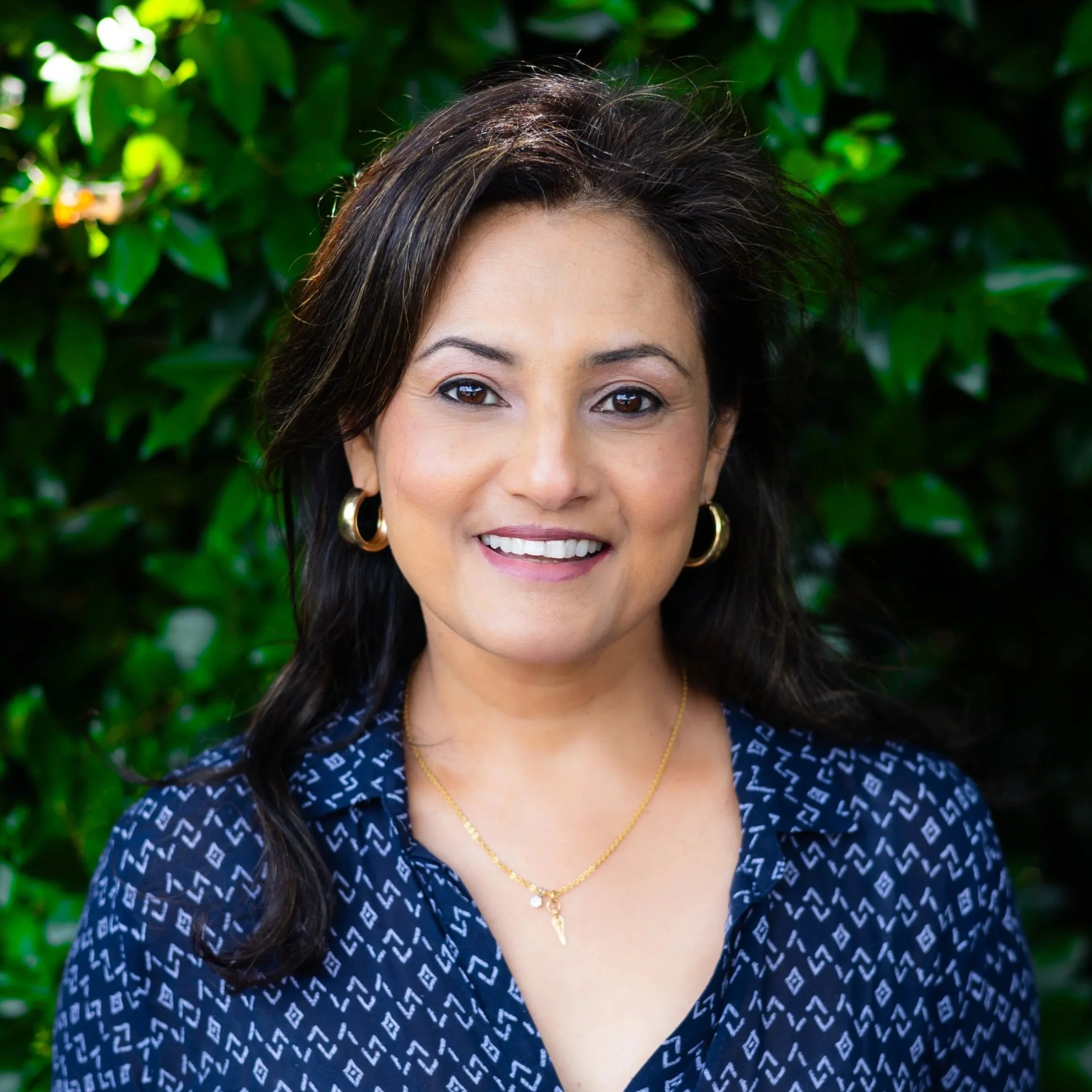 A woman with dark brown hair, wearing a blue patterned blouse, gold hoop earrings, and a gold necklace, smiling in front of a green leafy background.