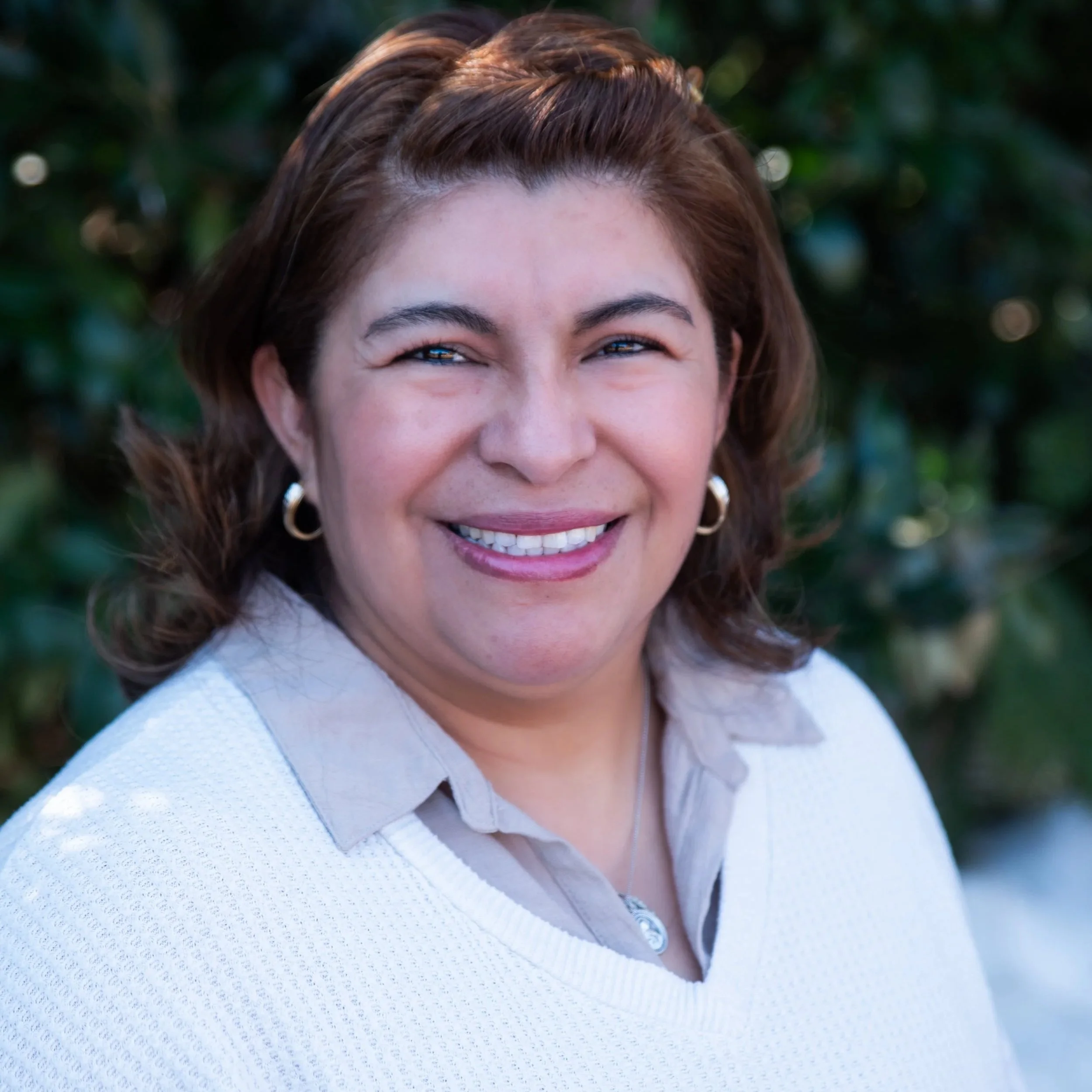 A woman with shoulder-length brown hair, smiling, wearing a light-colored collared shirt and earrings, outdoors with greenery in the background.