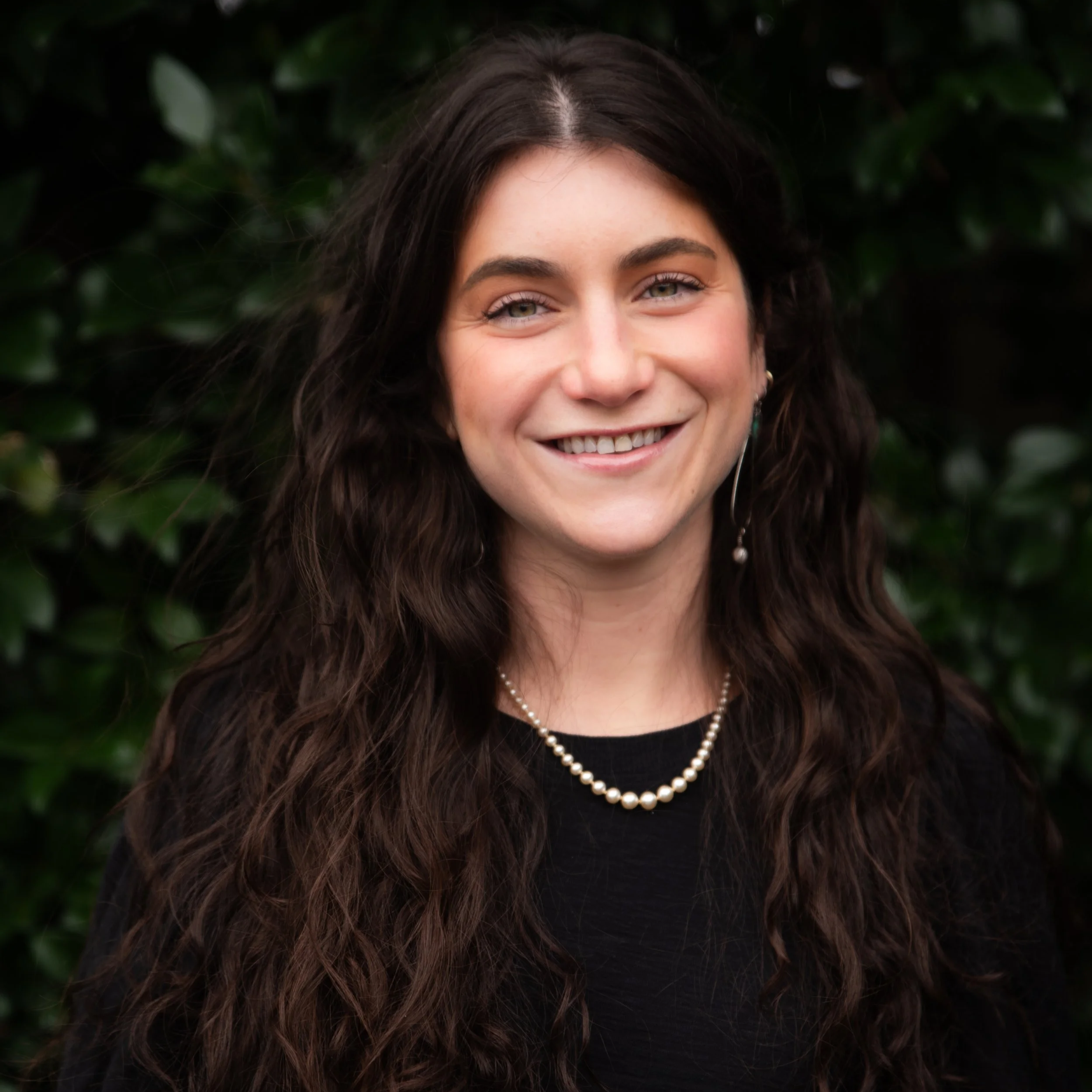 A woman with long curly dark brown hair, wearing a pearl necklace, earring, and a black top, smiling outdoors in front of green foliage.