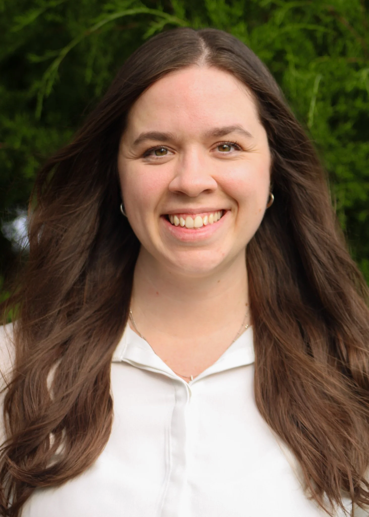 Portrait of a smiling woman with long brown hair, green eyes, wearing hoop earrings and a white collared shirt, outdoors with green foliage in the background.