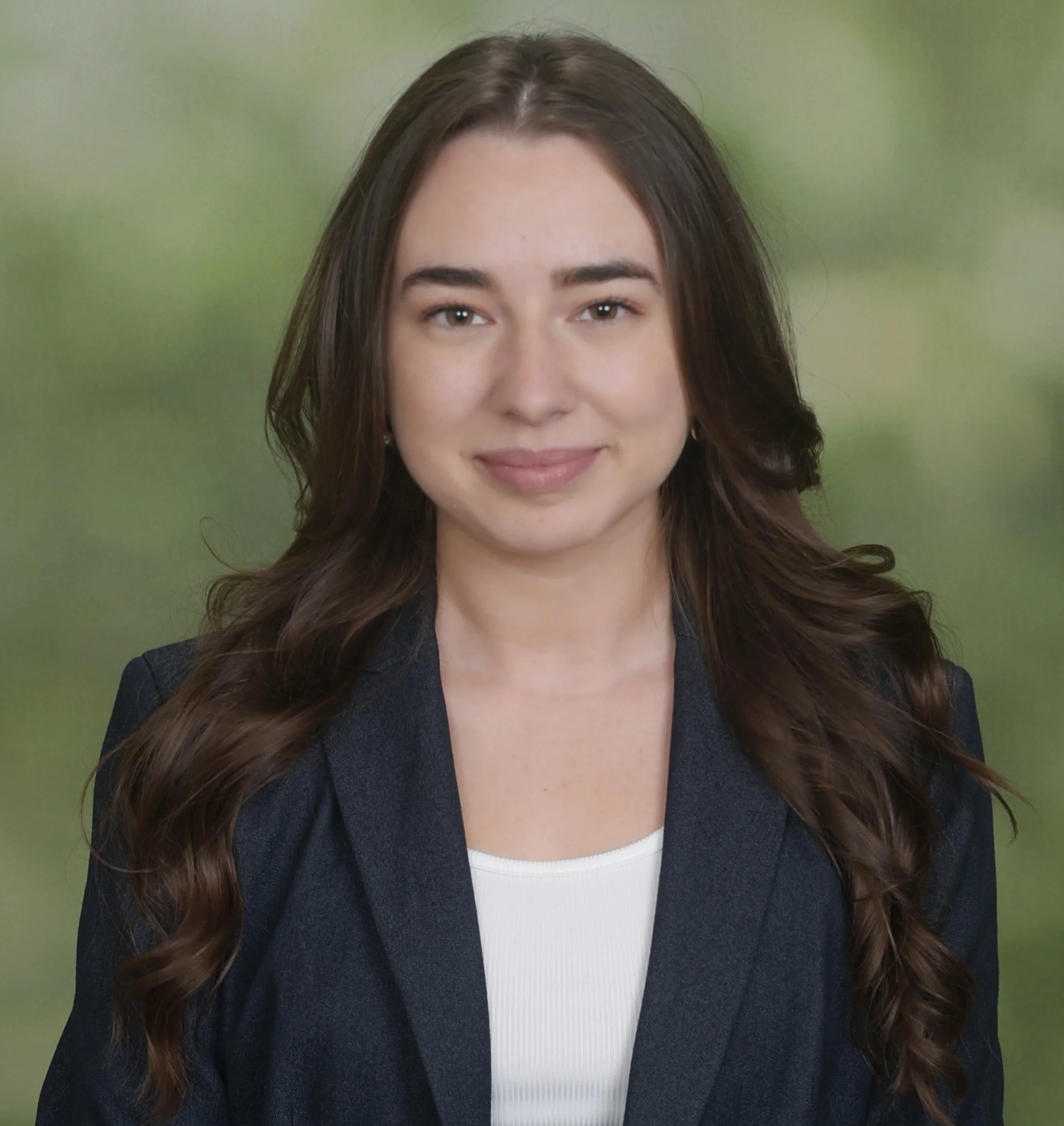 A young woman with long, wavy brown hair, wearing a dark blazer over a white top, smiling softly against a blurred green background.