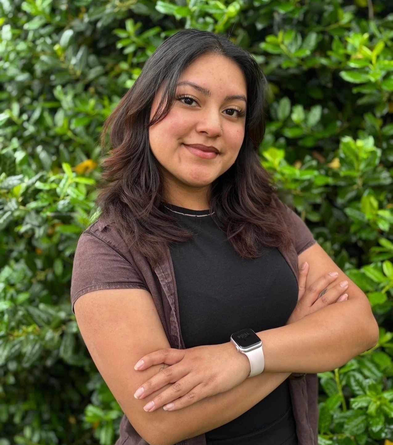 A young woman with shoulder-length dark hair, smiling and crossing her arms, standing in front of green foliage. She is wearing a black t-shirt, a brown jacket, and a white smartwatch.