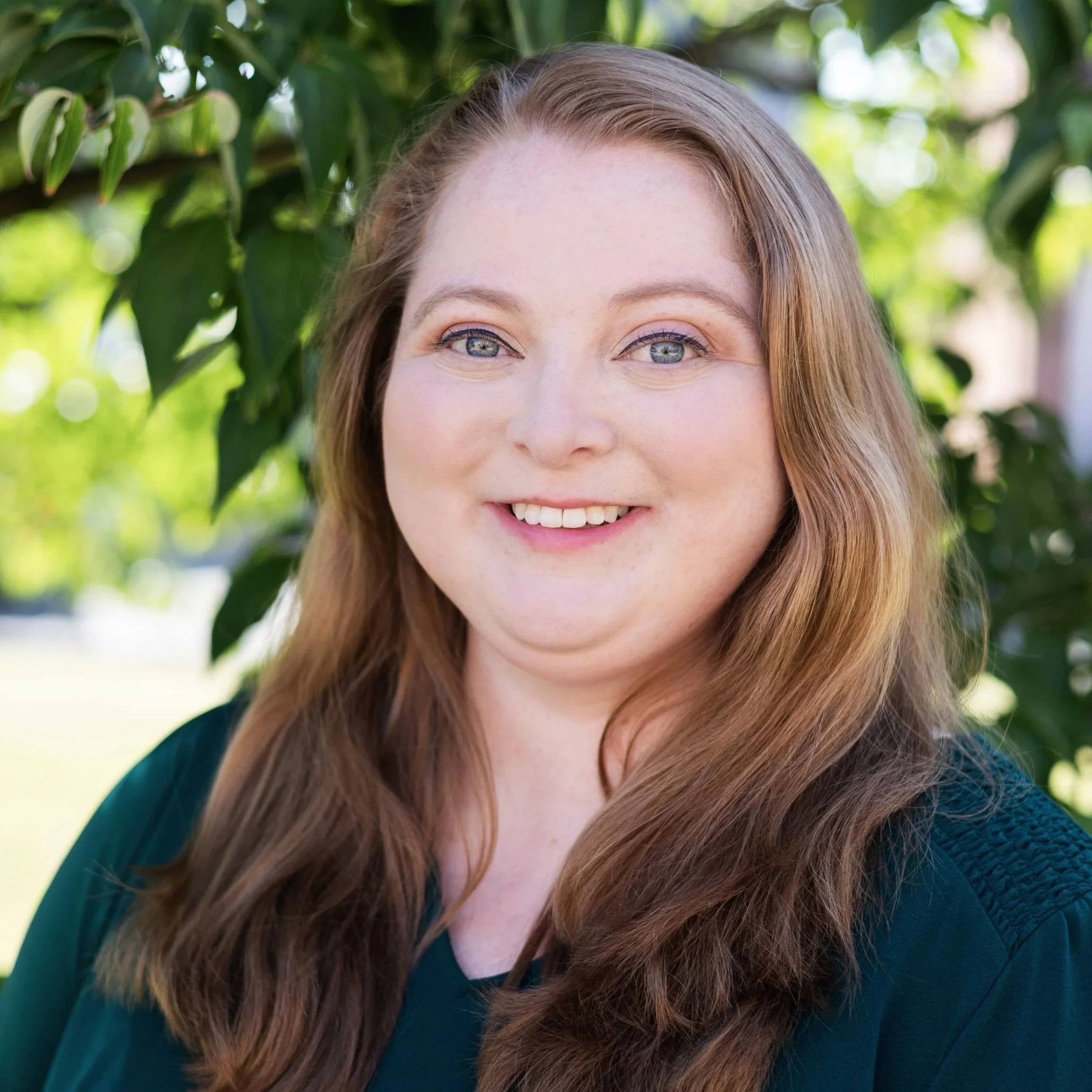Close-up of a woman with long, wavy auburn hair, smiling outdoors with green foliage in the background.